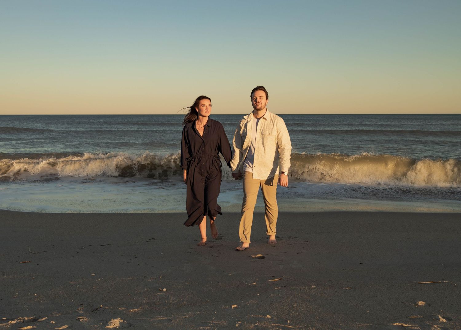 Couple walks hand in hand along shoreline during golden hour.