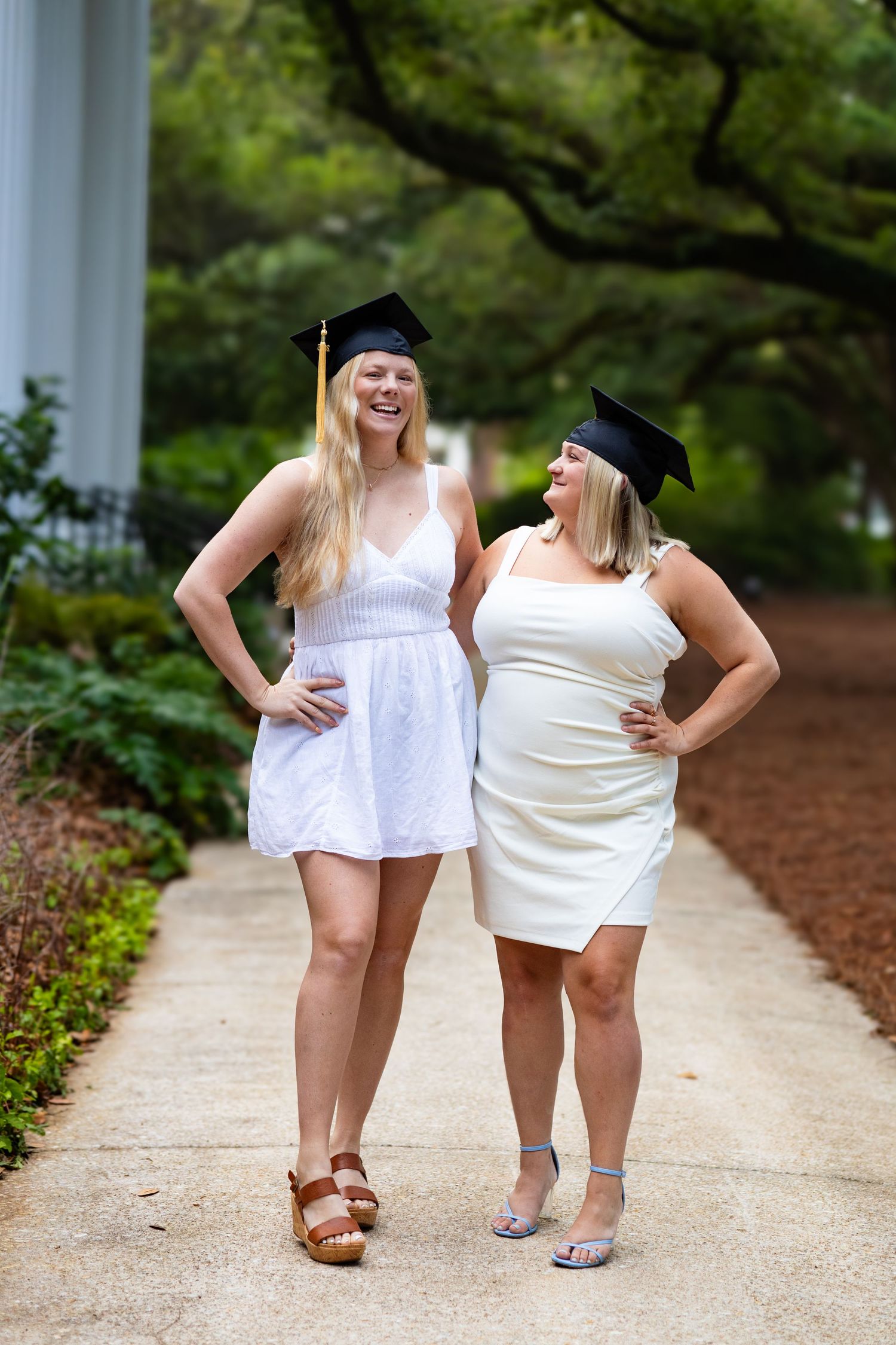 Two smiling female graduates in white dresses and black mortarboard caps pose together on a garden pathway.
