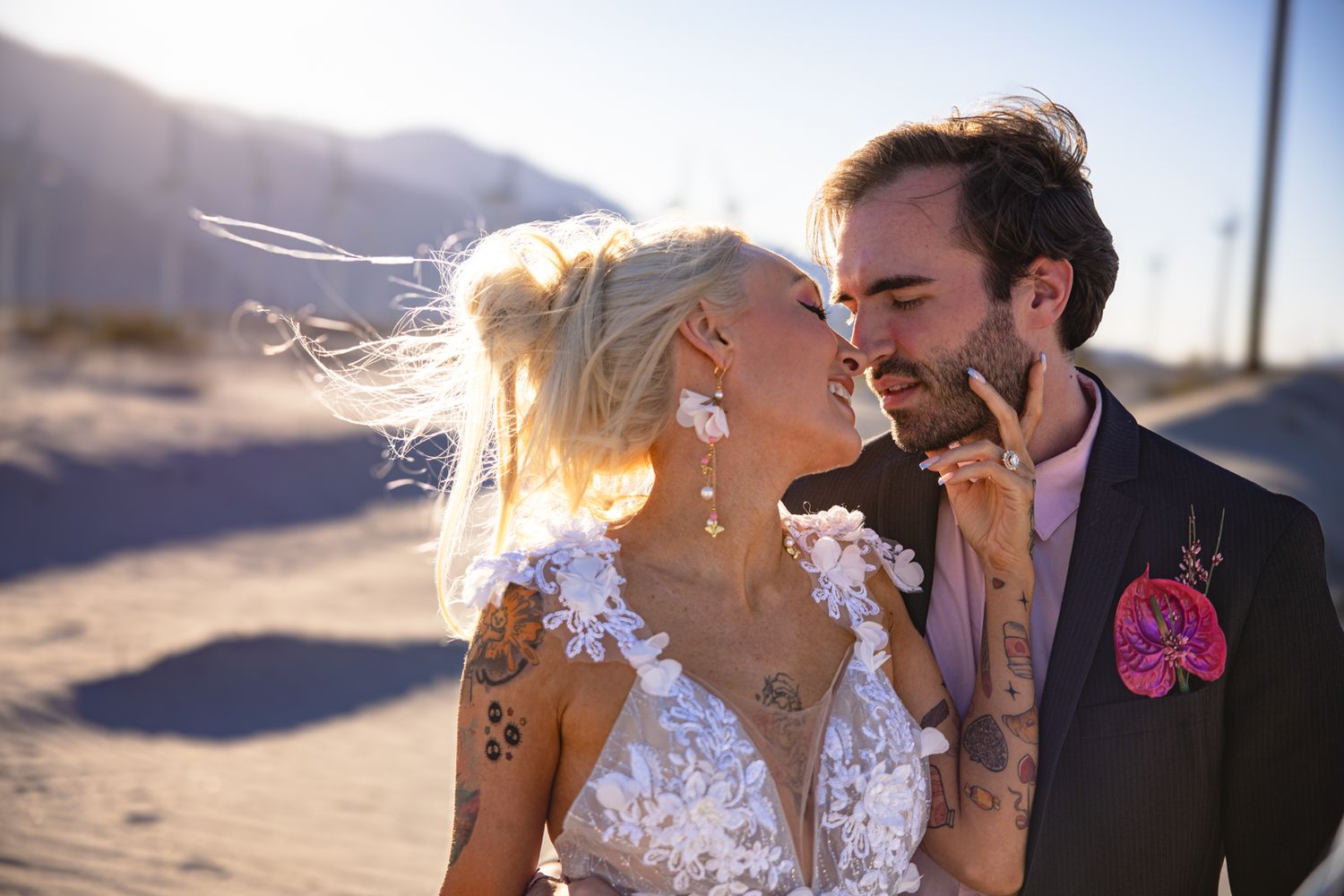 A bride and groom share a romantic moment on a desert beach, her lace dress and floral earrings complementing his pink suit.
