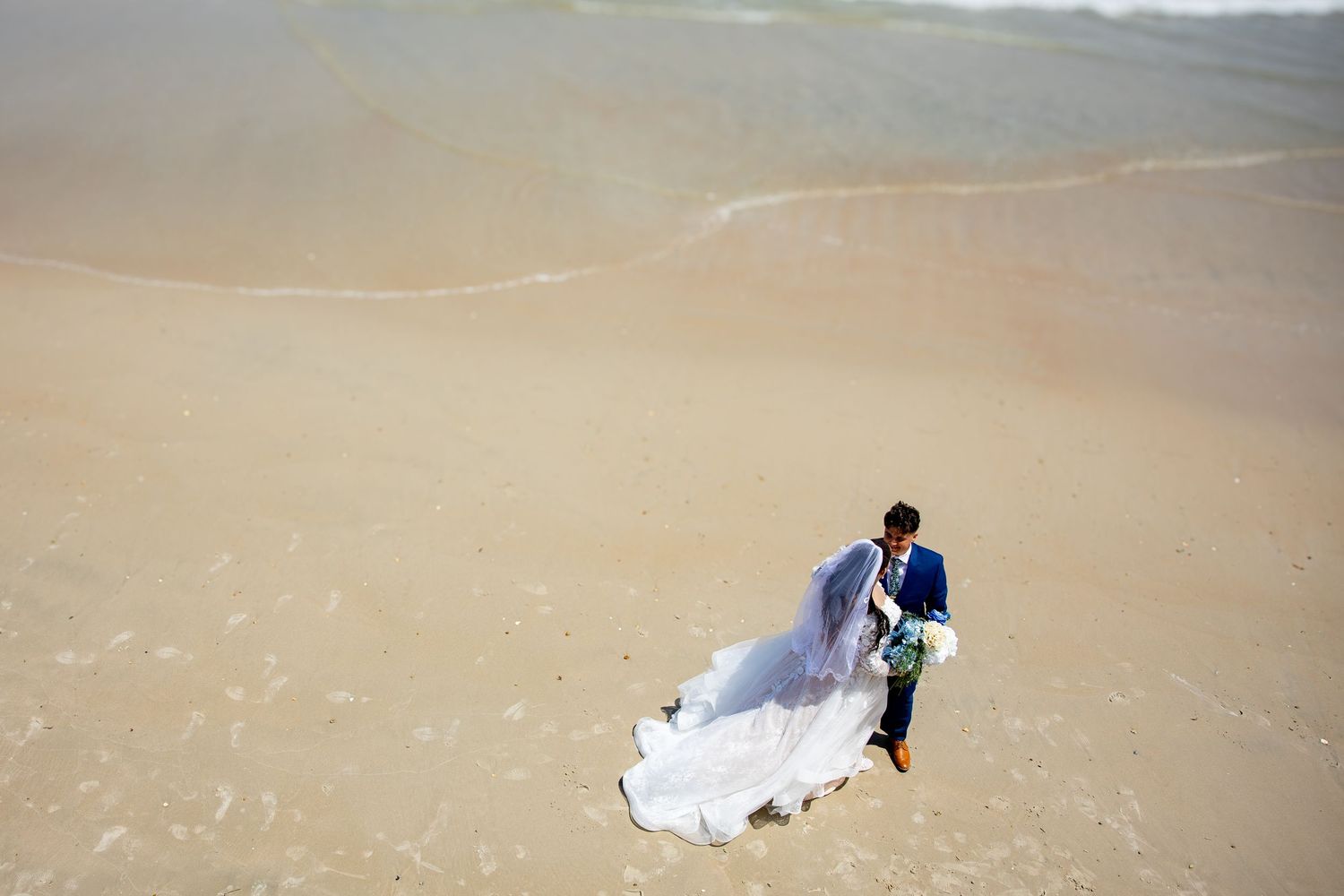 Bride and groom walking on sandy beach, wedding veil flowing in wind, aerial view of couple near ocean waves.