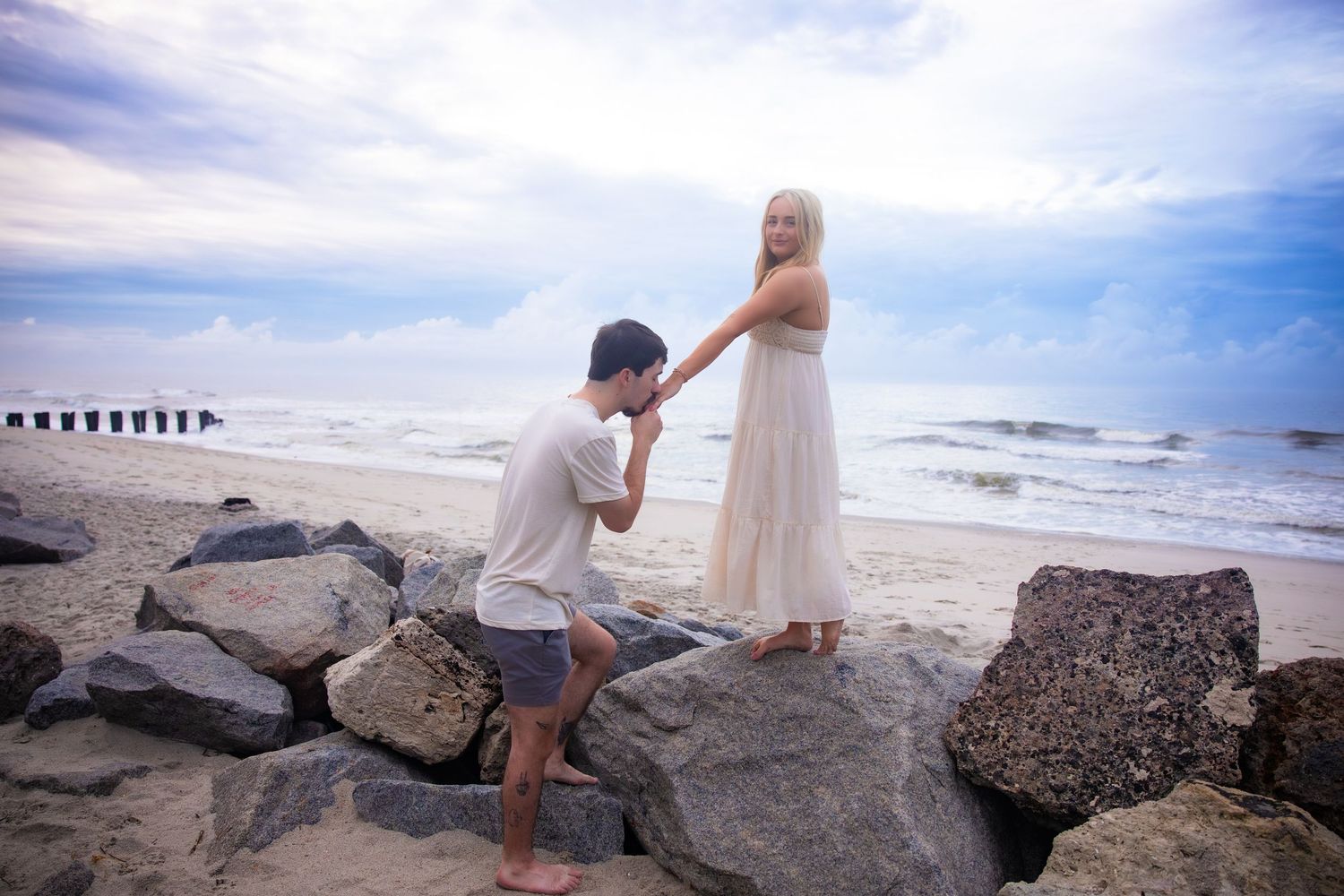 Romantic proposal moment on rocky beach with ocean pier in background.
