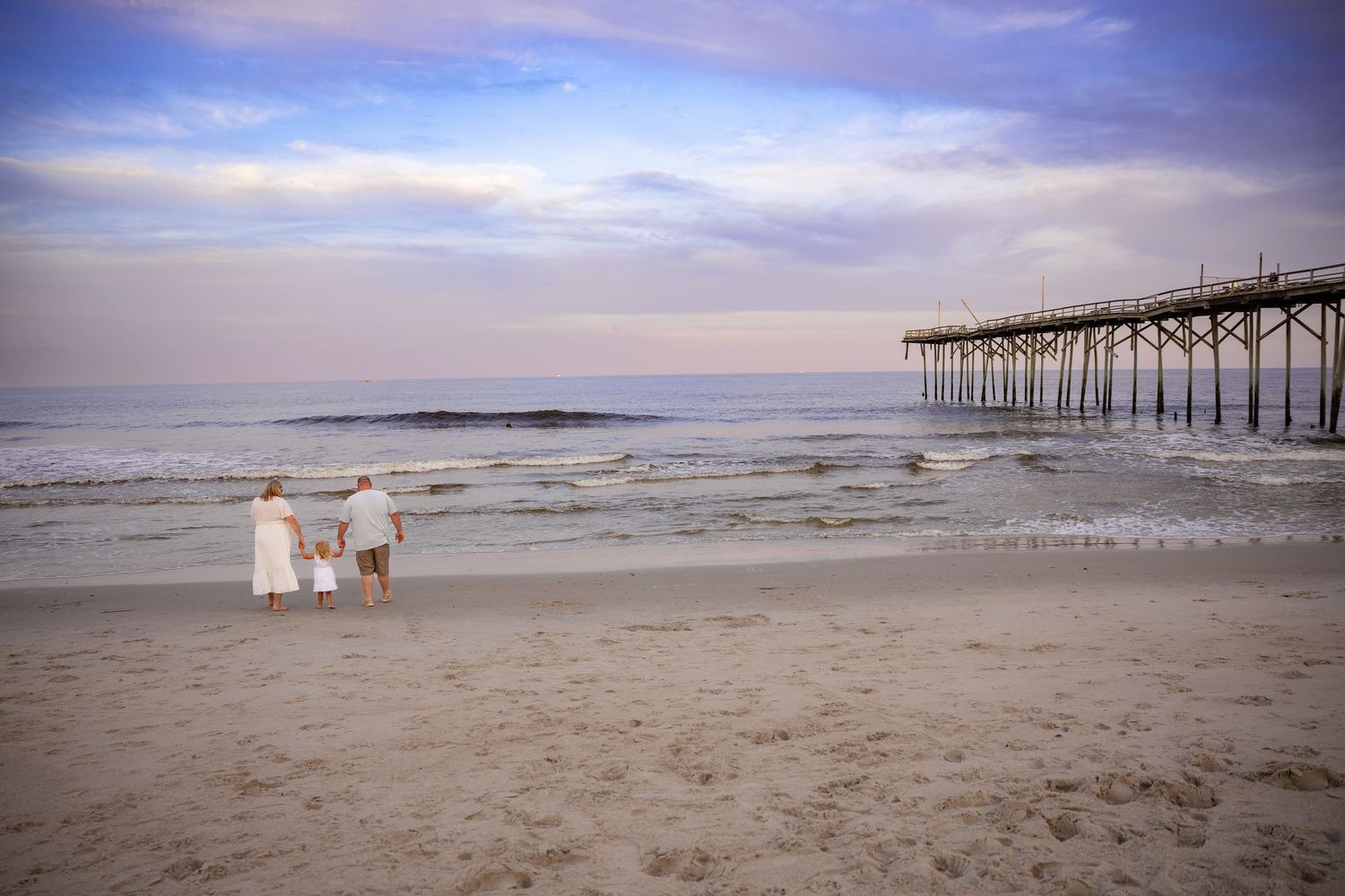 Two white chairs sit empty on a beach near a wooden pier at sunset.