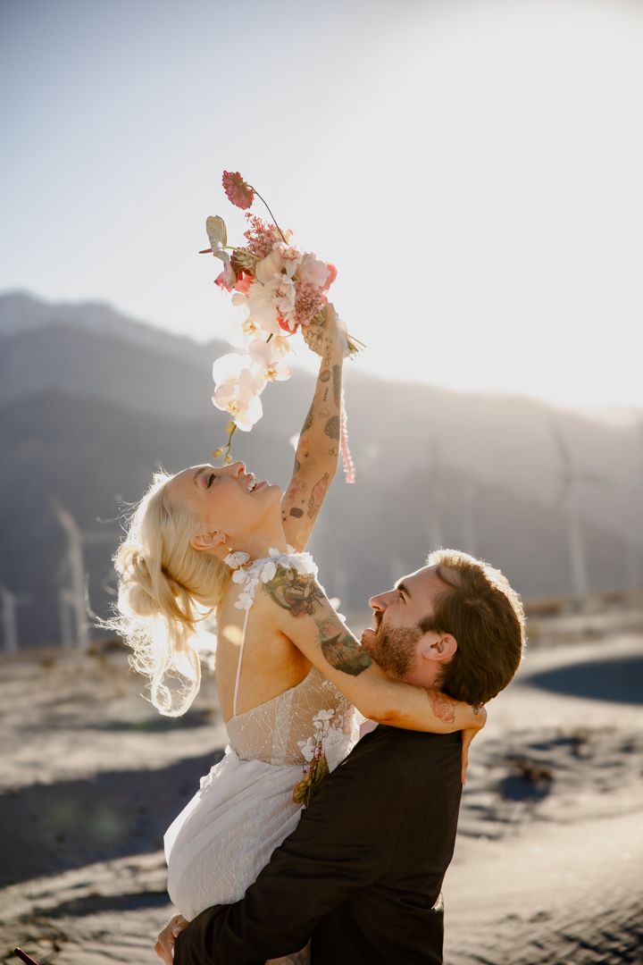 Groom lifting tattooed bride holding floral bouquet aloft against sunny mountain backdrop at desert wedding.