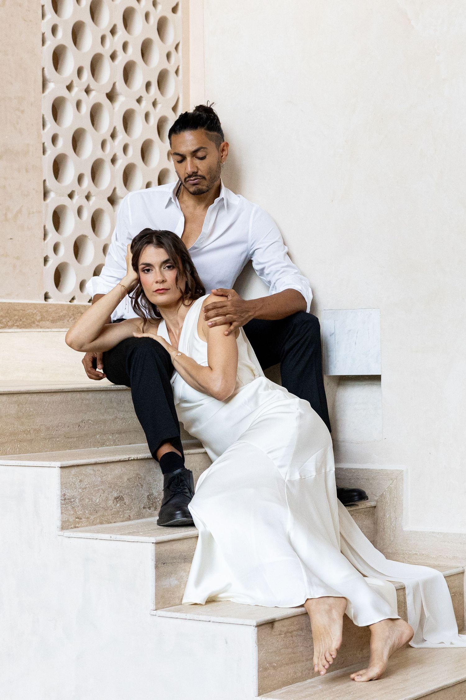 Elegant couple posing on marble steps, woman in white silk dress, man in white shirt and black pants.