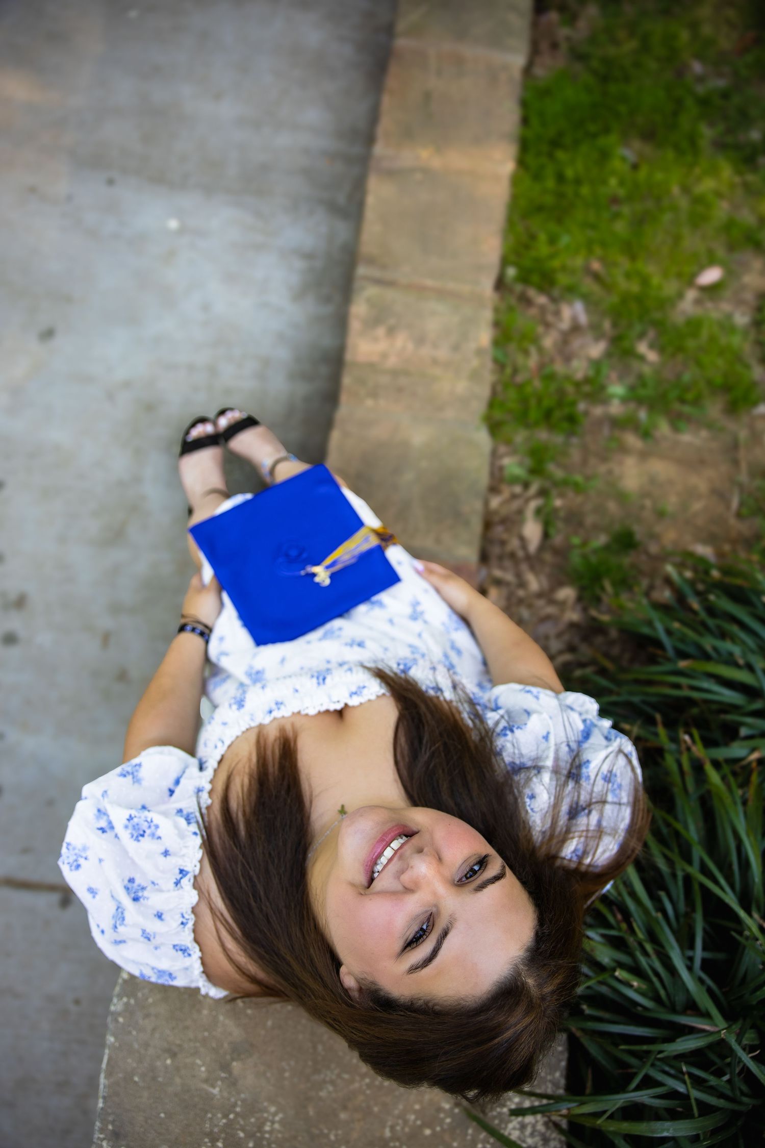 Young woman in floral dress lying on steps outdoors, smiling up at camera while holding a blue book.