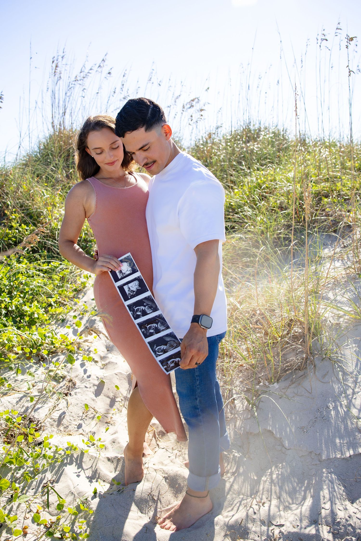 A couple embraces on a sunny beach with sea grass and sand dunes in the background.