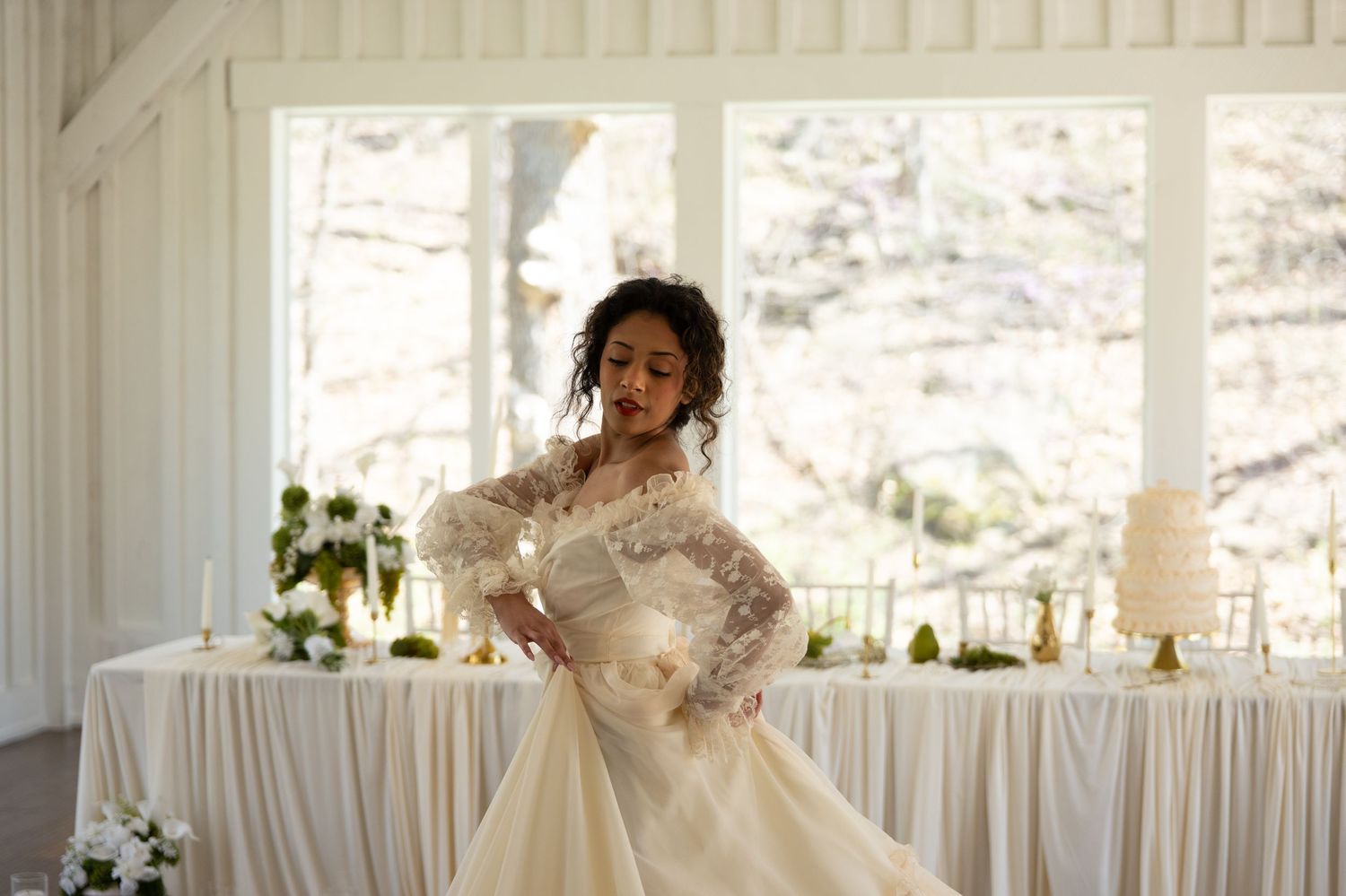 Bride in elegant lace long-sleeve wedding gown twirling in a bright venue with floral decor and a tiered cake.