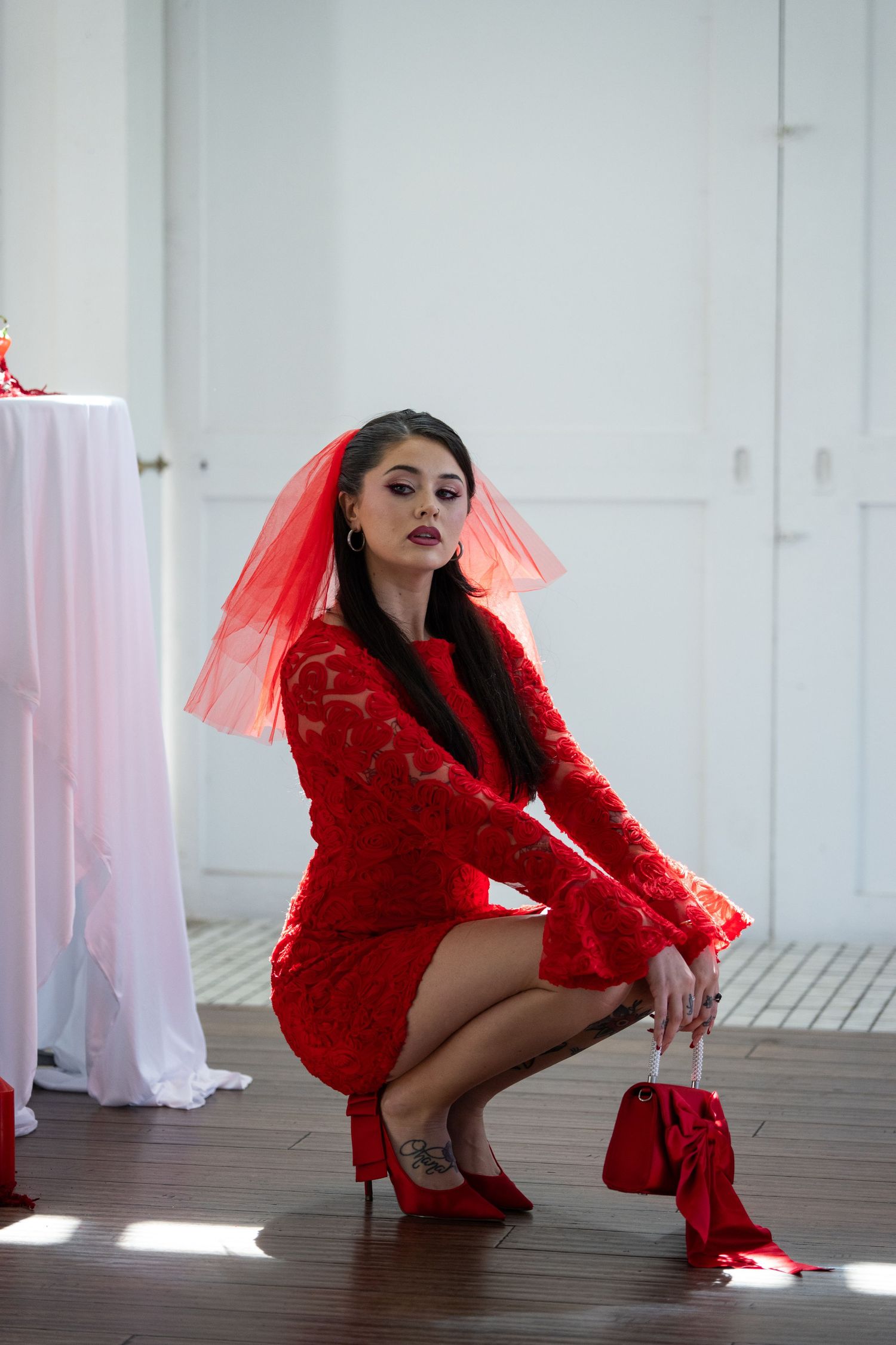 Woman in red lace dress and red veil crouches gracefully on wooden floor holding a small red handbag.