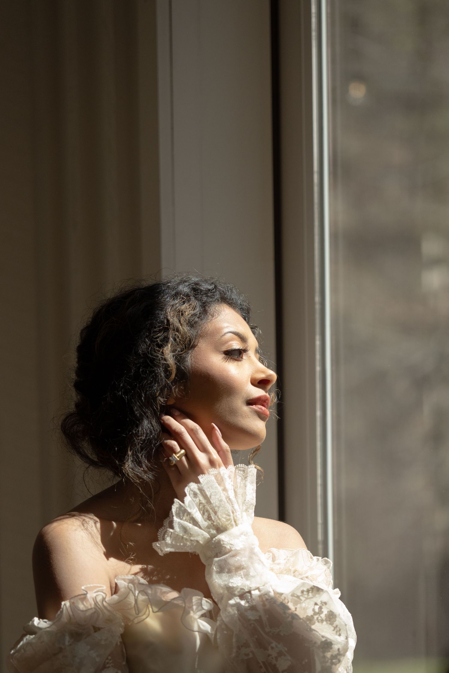A woman in a white lace off-shoulder dress gazes out a sunlit window, hand gracefully touching her neck.