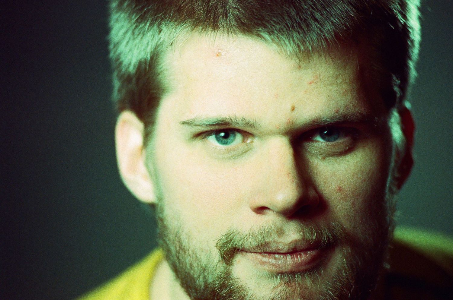 Close-up portrait of a young man with blue eyes, short hair, and a beard, lit dramatically against a dark background.