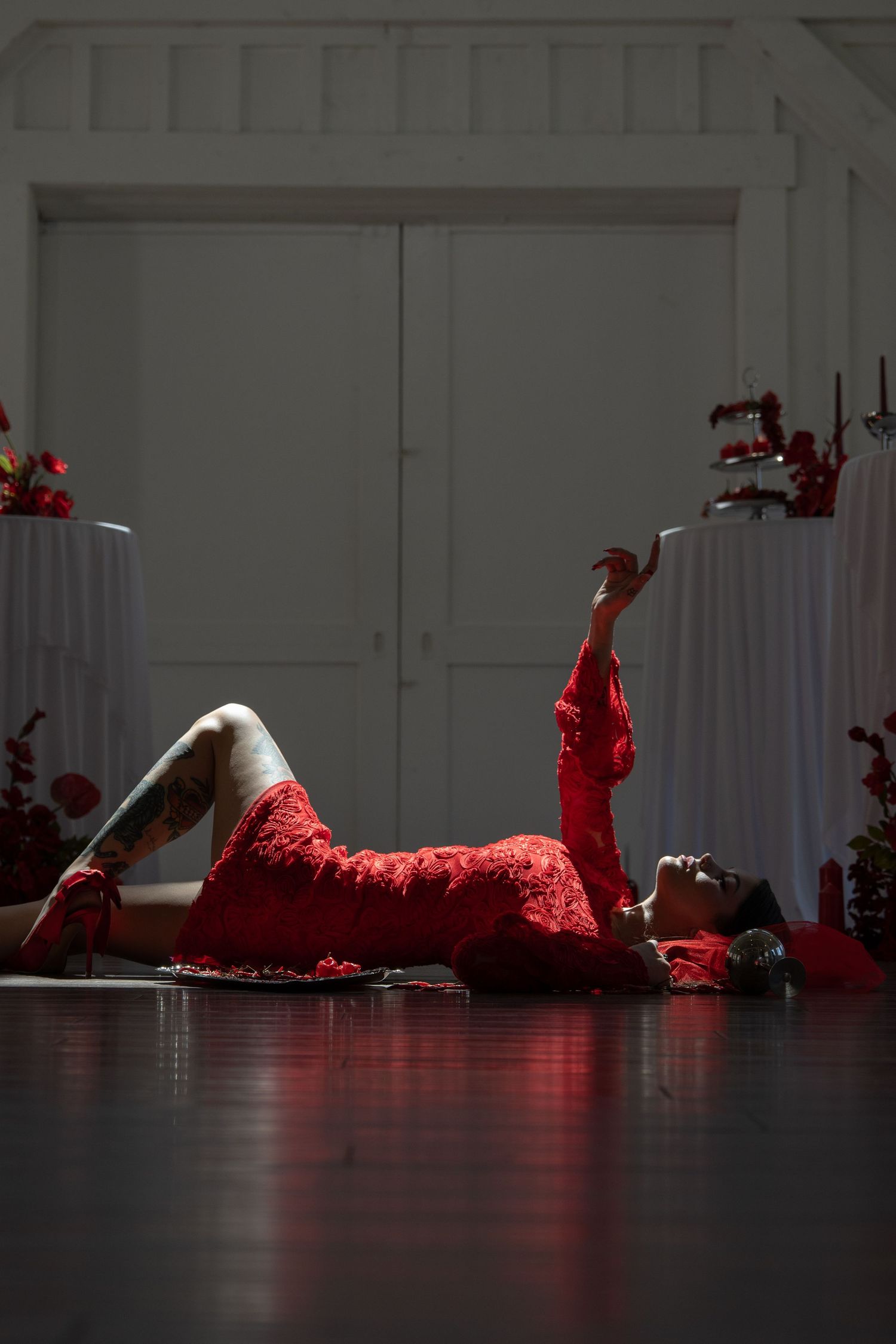 Woman in red lace dress lying on dark floor, posing elegantly with arm raised in dramatic indoor setting.