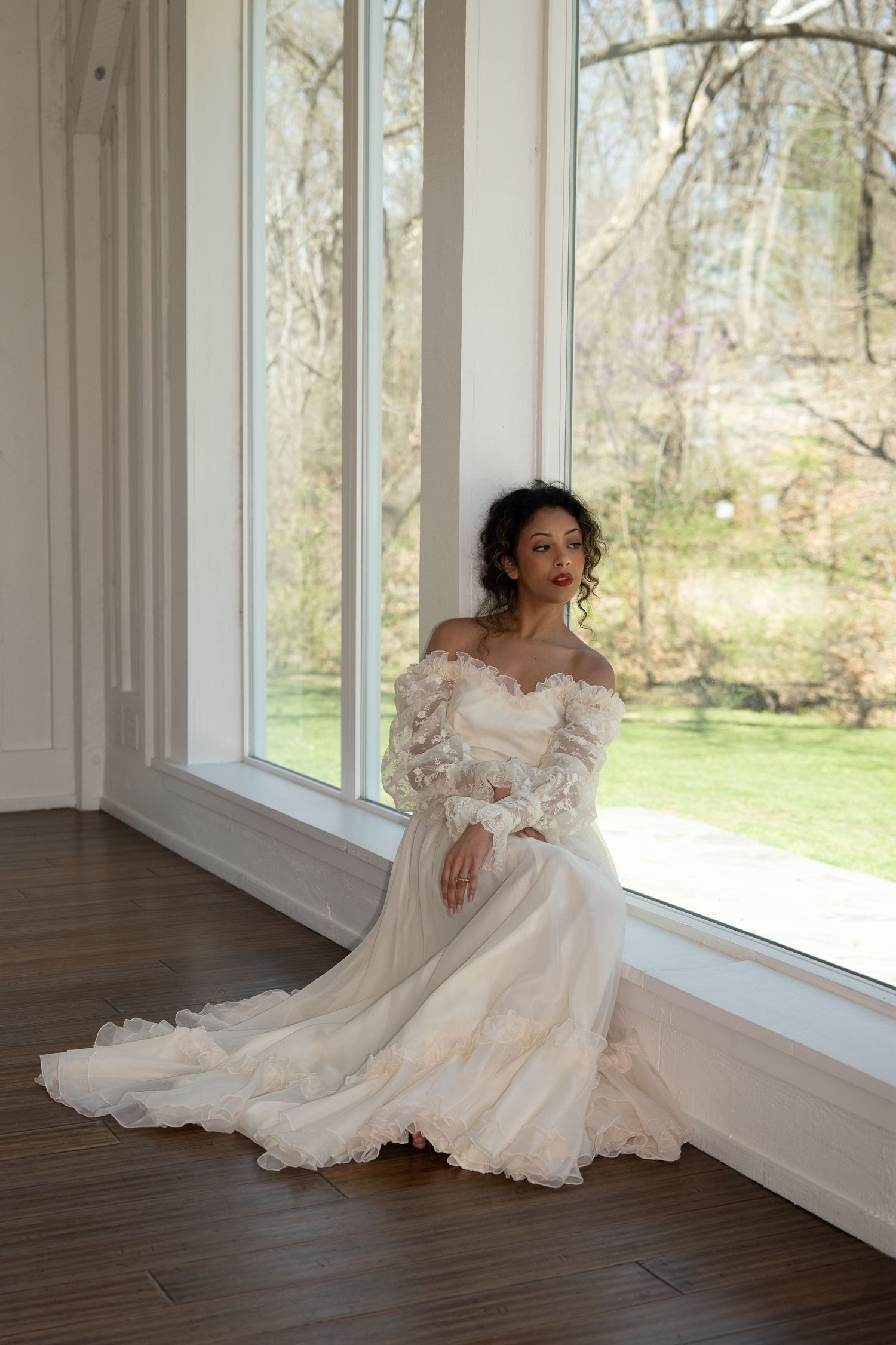 Bride in off-shoulder lace wedding gown with floral details sits gracefully on floor by large windows.