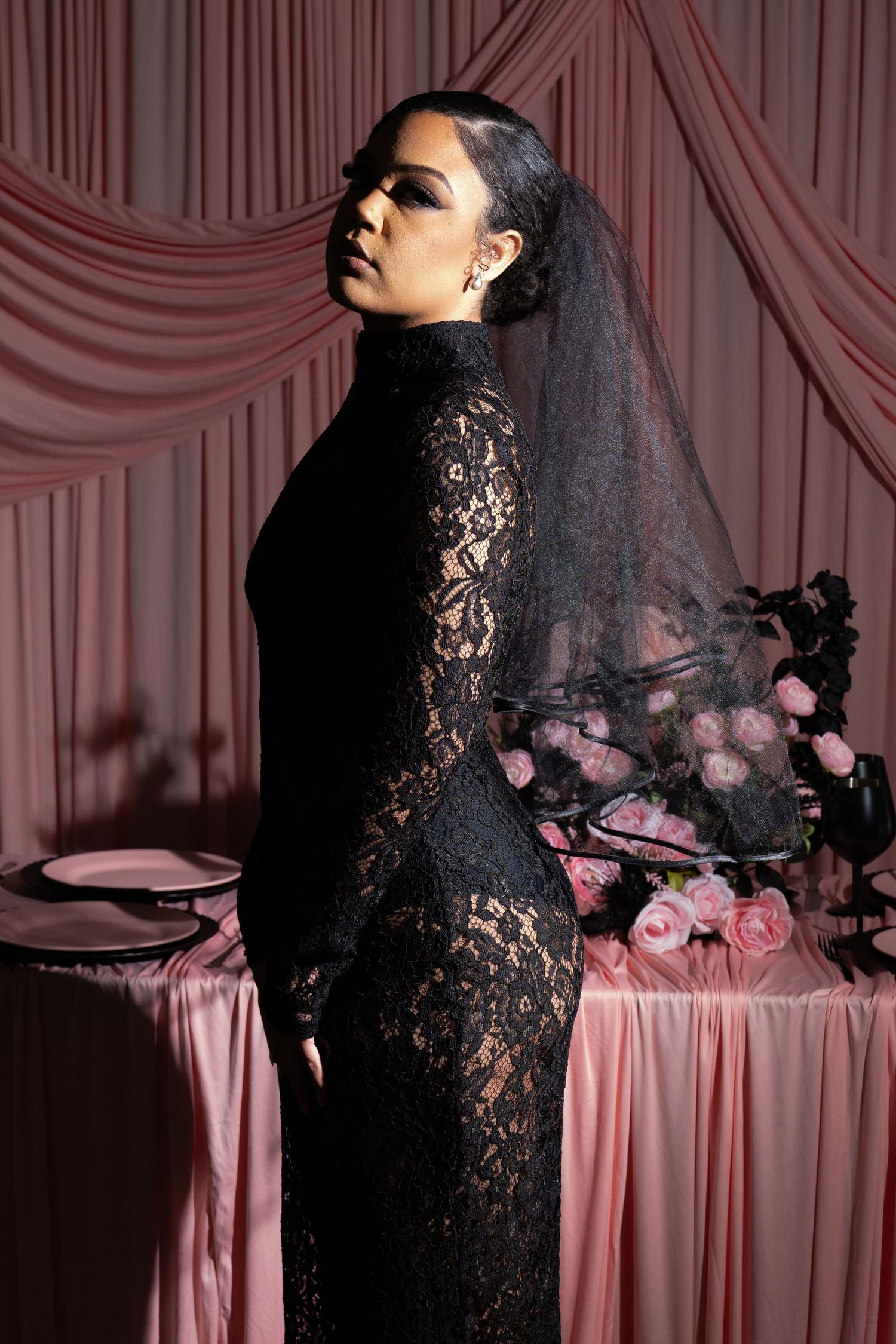 Woman in black lace dress and veil poses elegantly beside a pink draped table with roses.