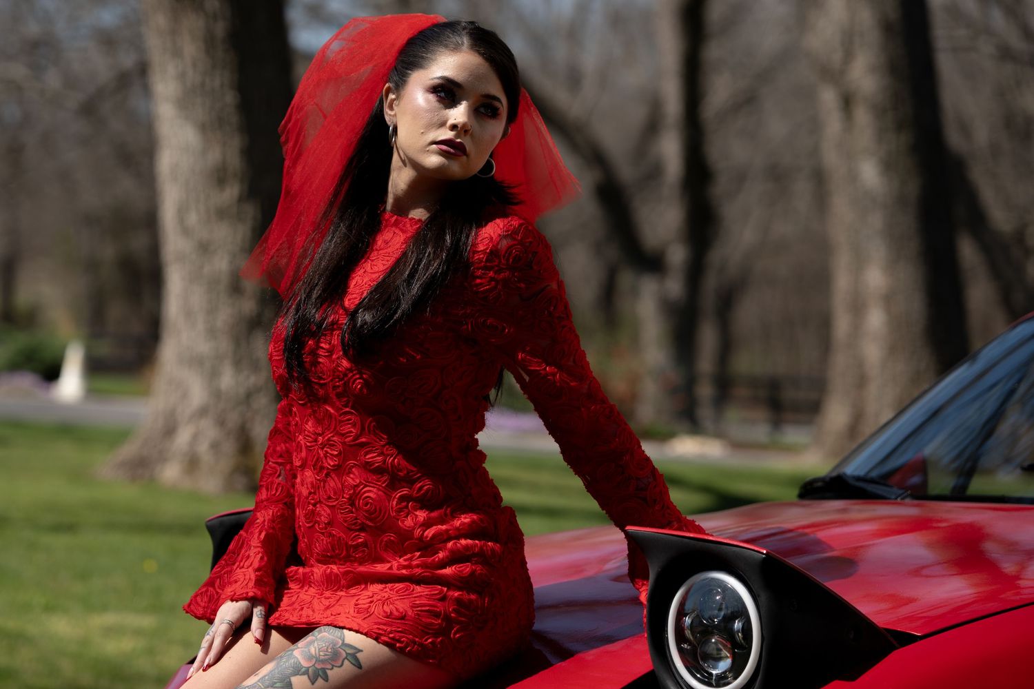 Woman in red lace dress and red veil poses on red sports car in a park setting.
