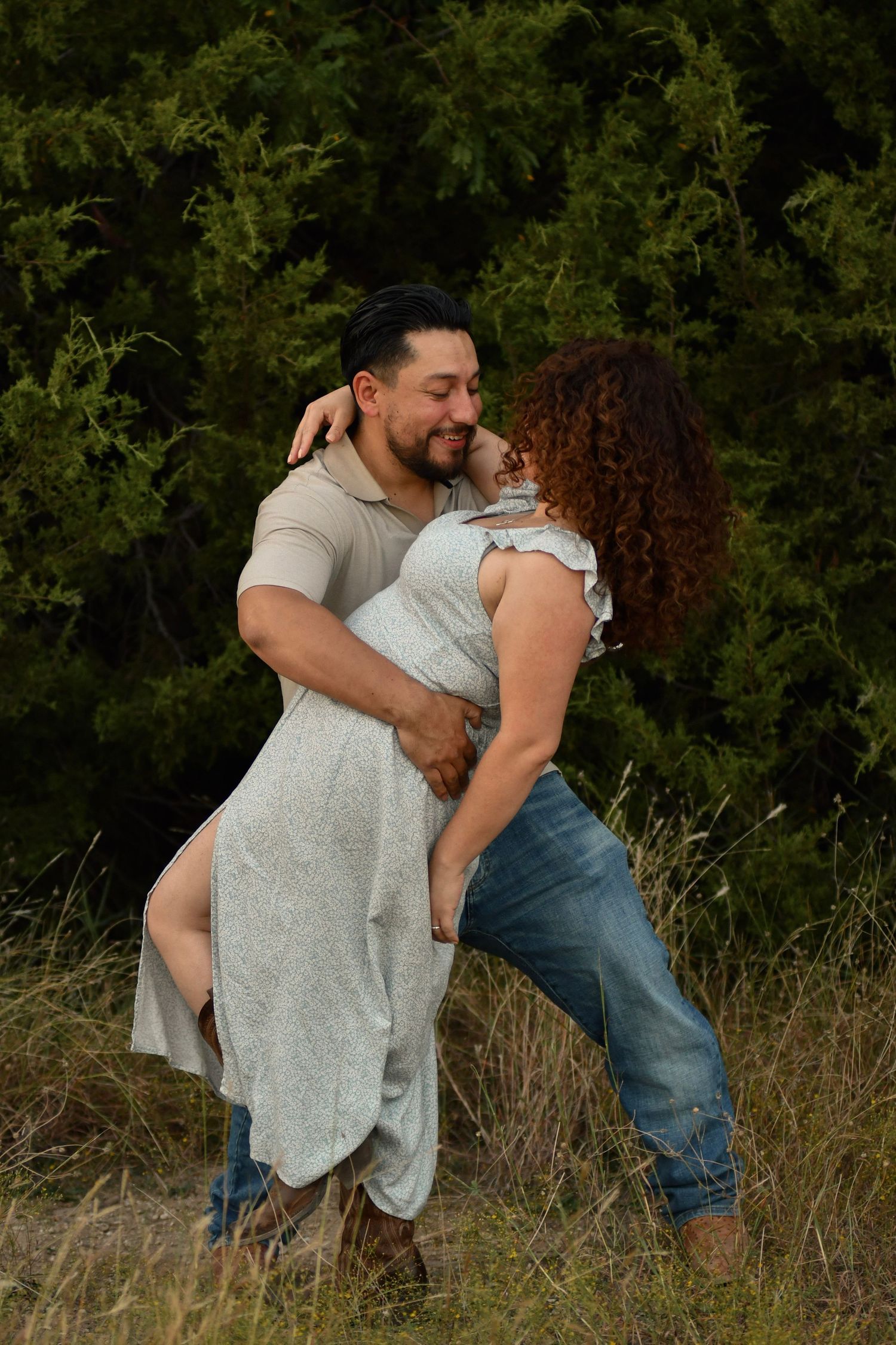 A playful couple embraces and dances together in a grassy outdoor field at sunset.