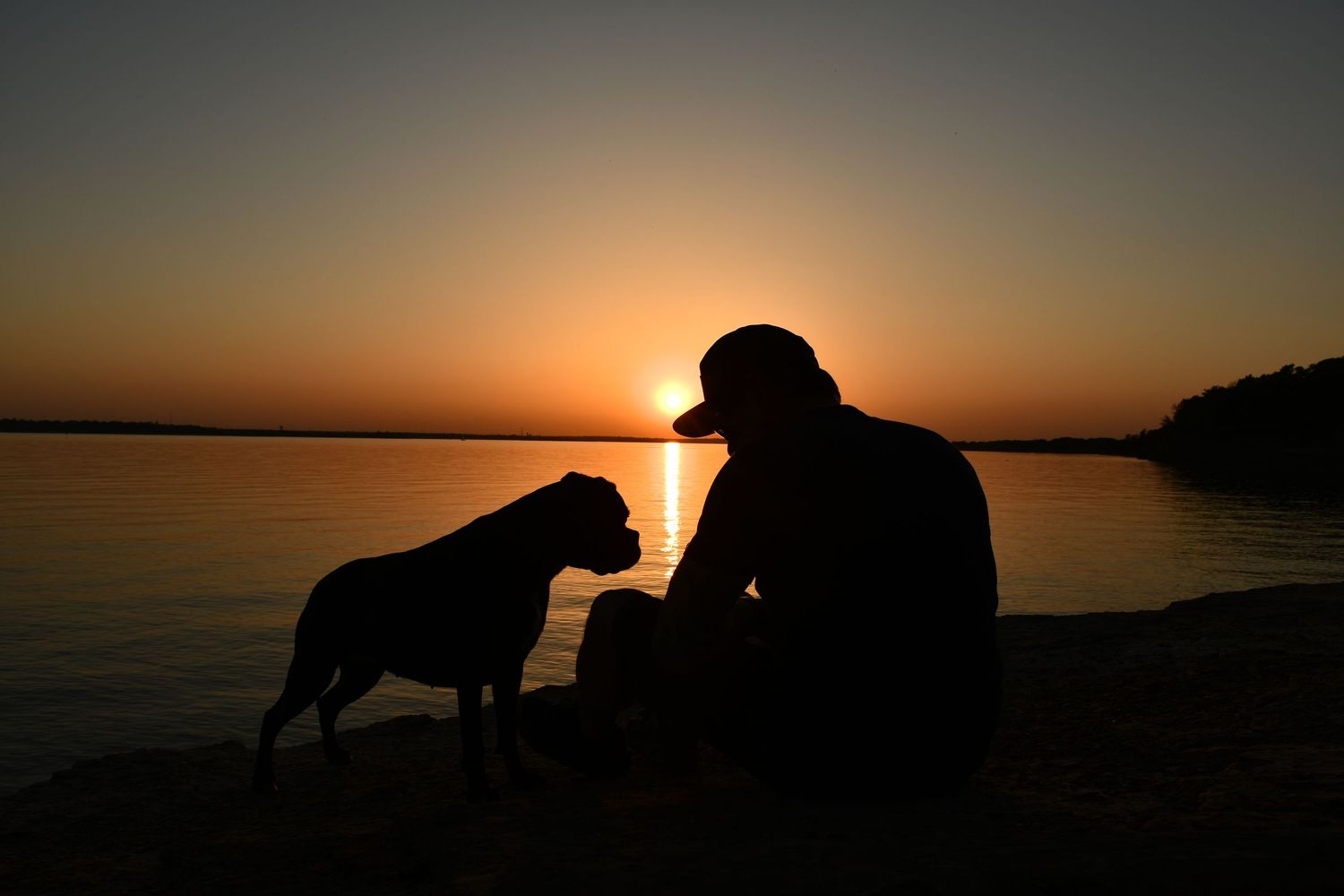 Texas photographer captures silhouette of handsome man and pet dog against dramatic sunset on lake shore.