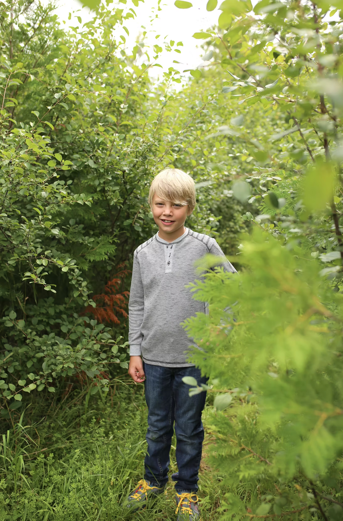 Young child in grey sweater and jeans exploring lush green foliage in garden setting.