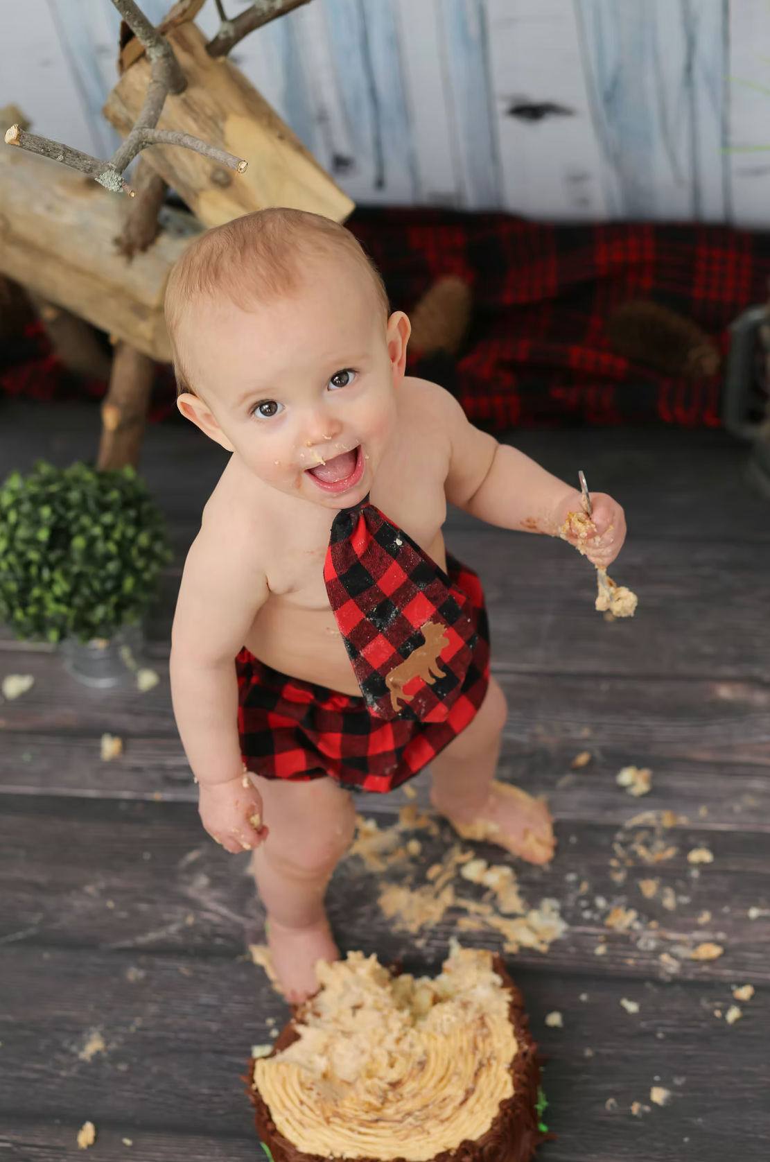 Baby in red plaid outfit plays with cake smash setup on rustic wooden floor.