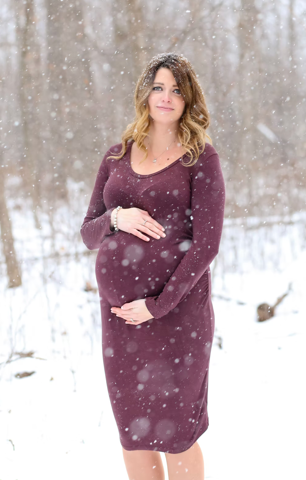 A maternity photo captures falling snow around a woman in a burgundy dress.