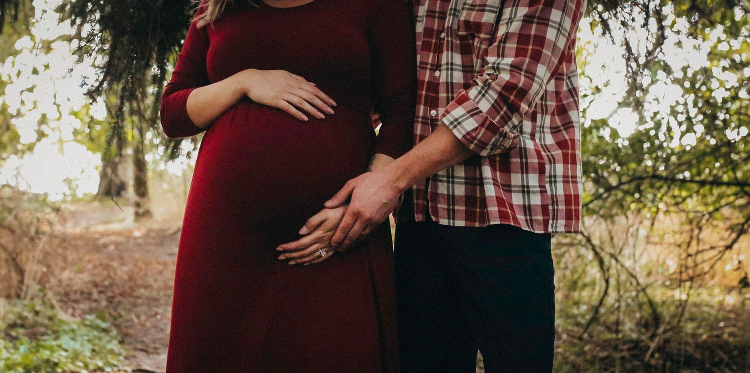 A couple poses together in a wooded outdoor setting wearing burgundy and plaid.