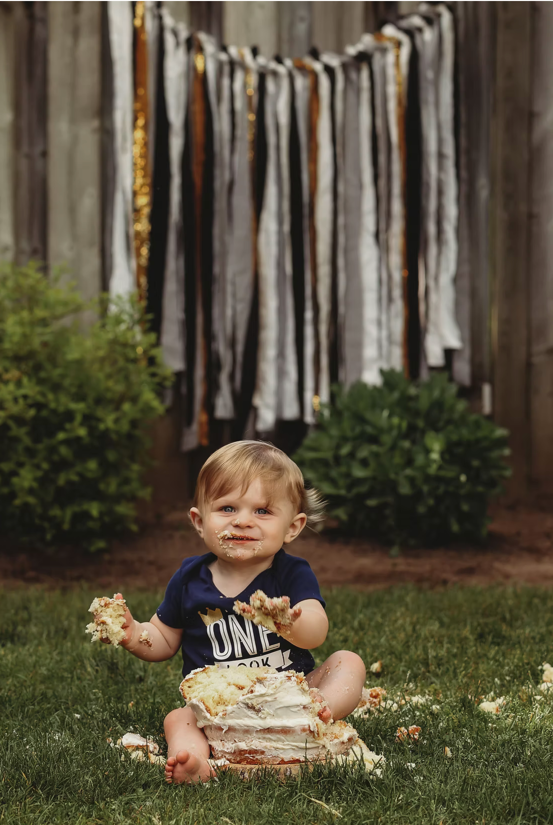 Rustic outdoor cake smash setup with black and gold fabric streamers and green foliage.