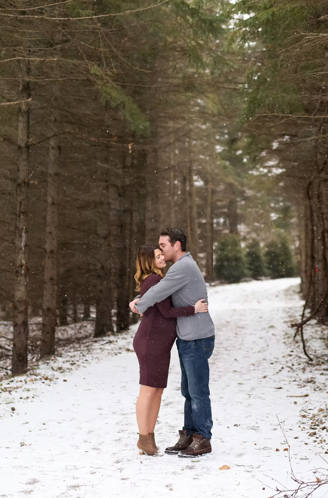 A romantic winter scene shows a couple embracing on a snow-covered forest path.