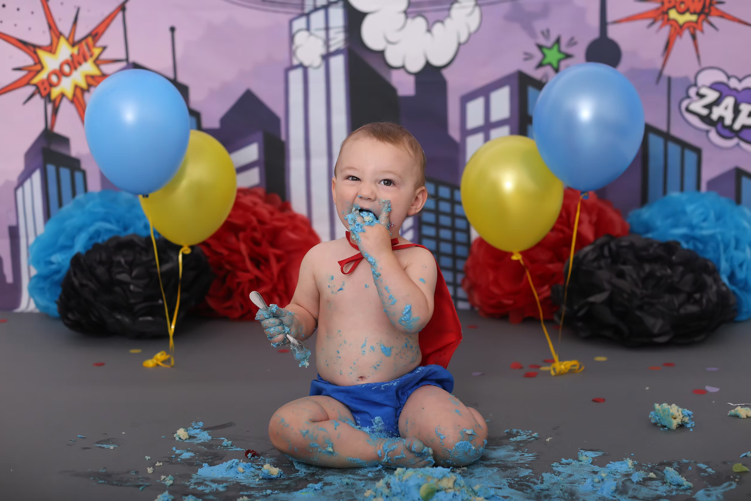 Festive superhero themed cake smash setup with blue and yellow balloons against comic backdrop.