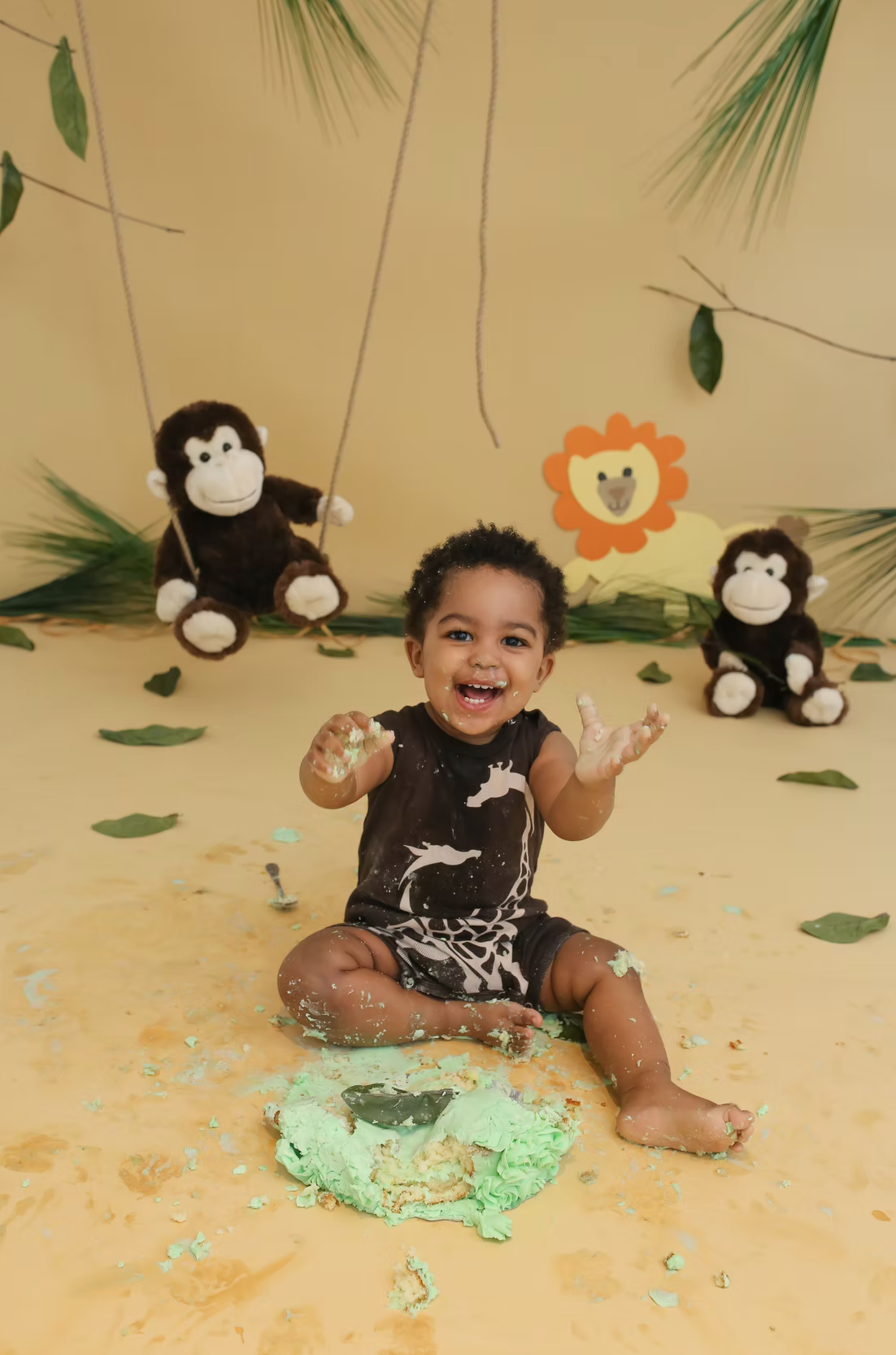 Cake smash photo session with green frosting and playful monkey plush toys against beige backdrop.