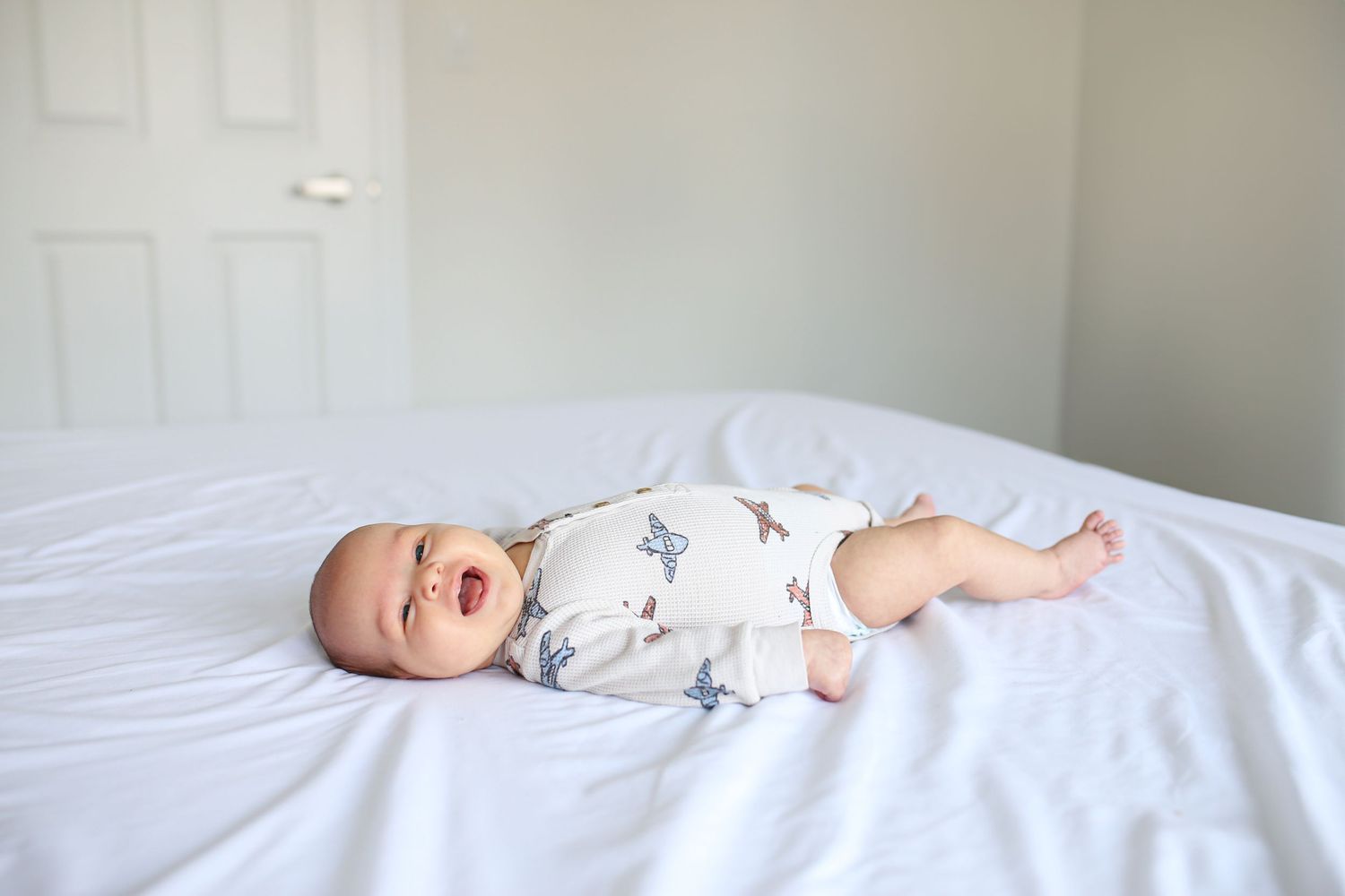Baby in white onesie lying on crisp white bedding in natural light.