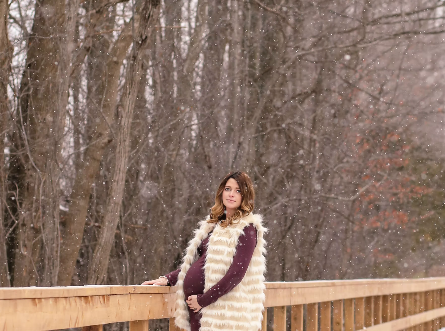 A maternity photo taken on a wooden bridge surrounded by winter trees.