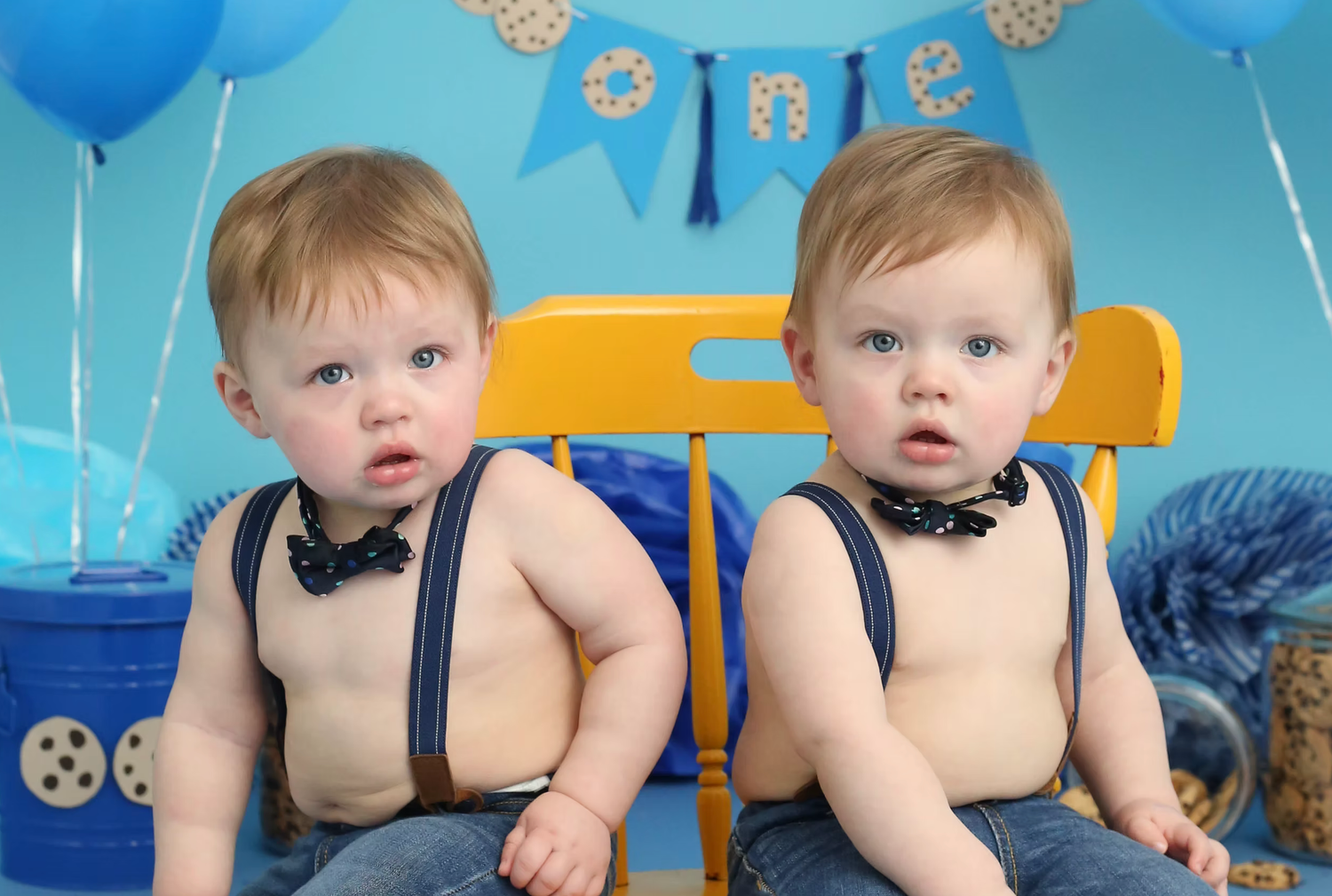 Twin boys in matching suspender outfits against blue party backdrop.