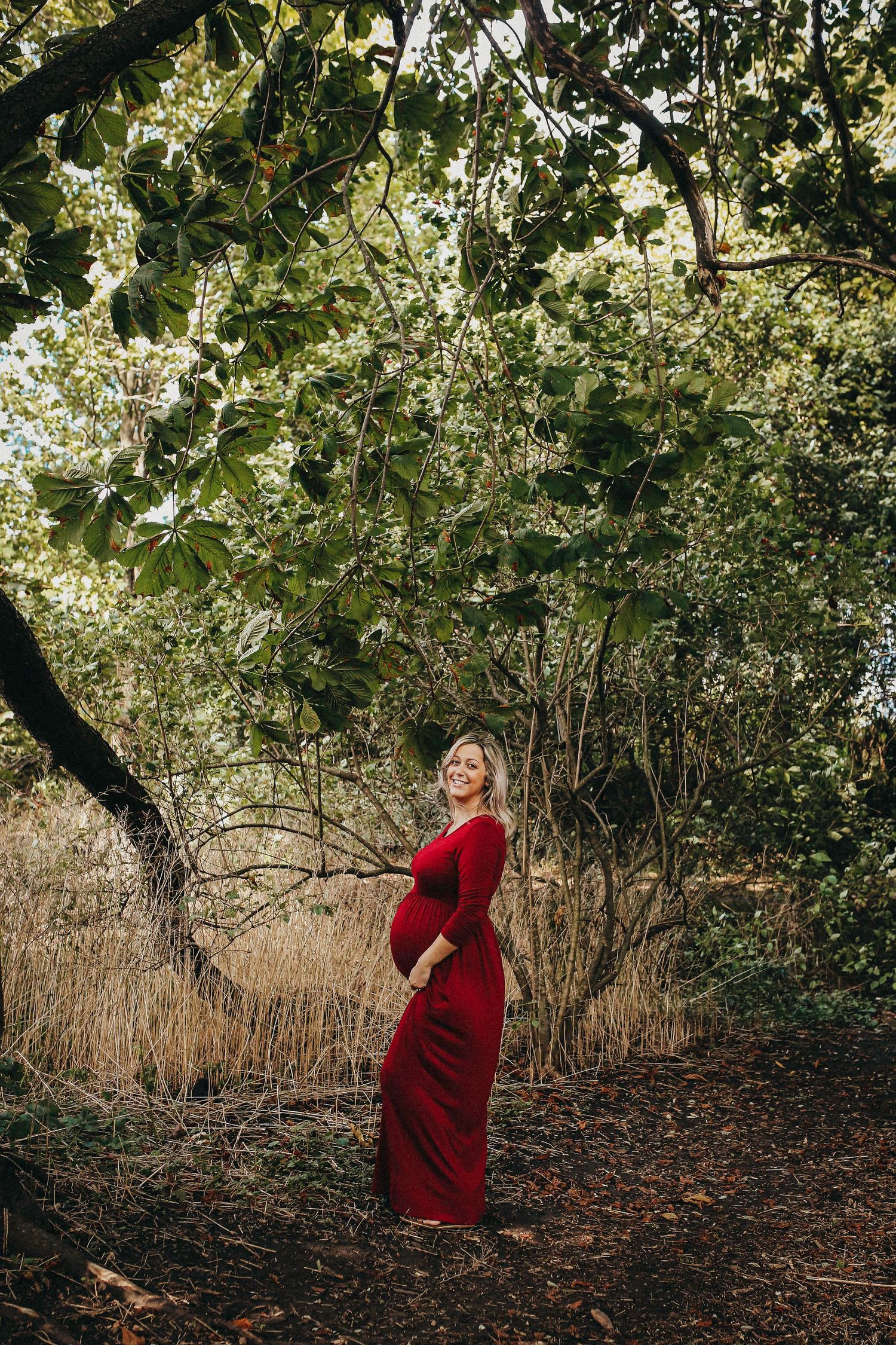 Woman in burgundy dress poses among trees and autumn foliage during golden hour.
