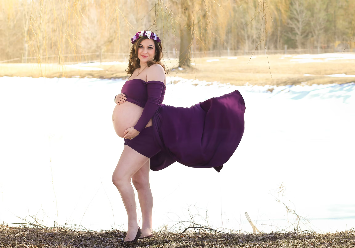 A flowing purple maternity dress billows in the breeze during an outdoor photo session near a lake.