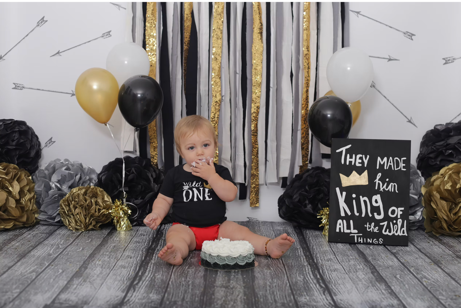 First birthday cake smash setup with gold and black balloons against rustic backdrop.