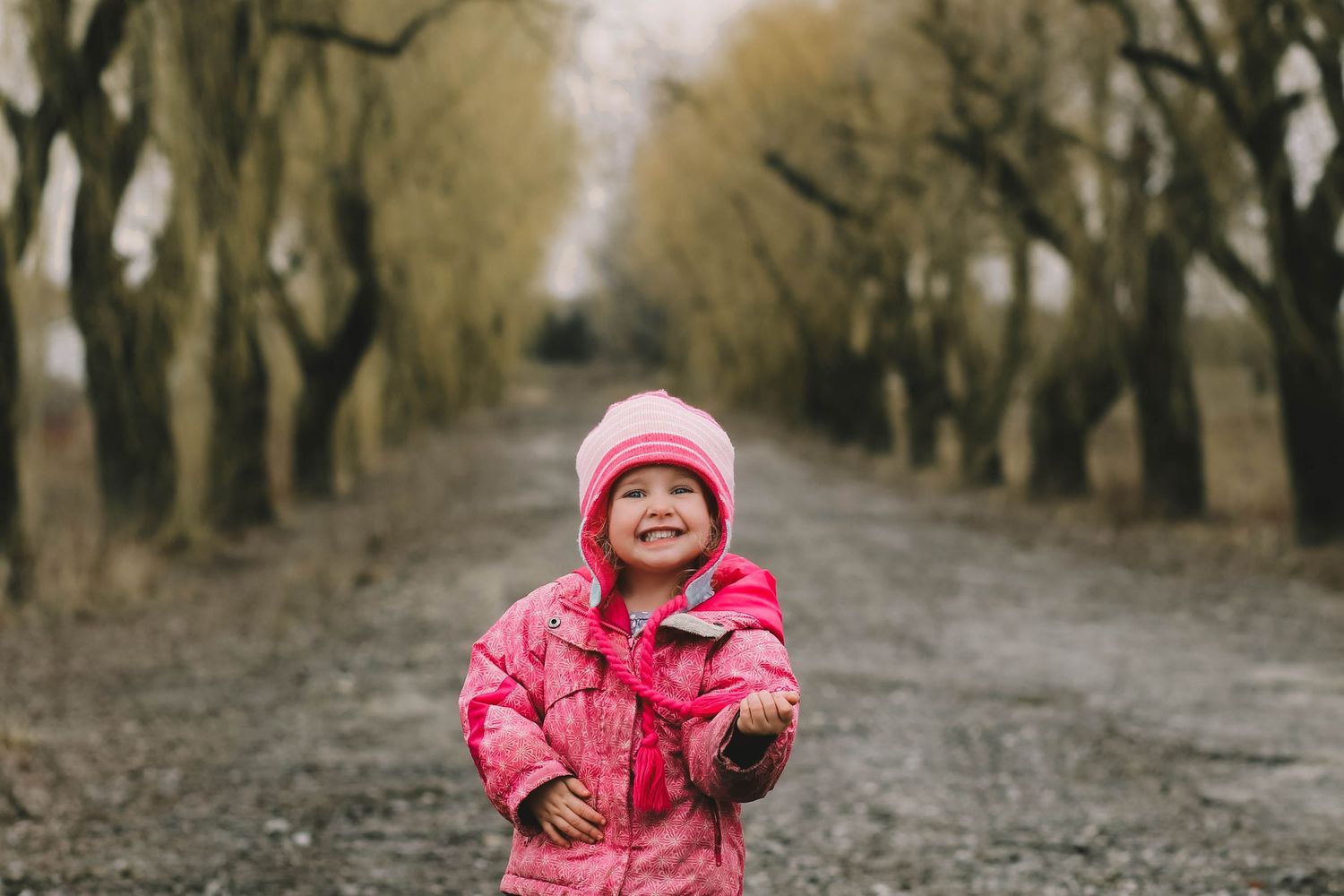 Child in bright pink winter coat walking along a tree-lined path on a moody autumn day.