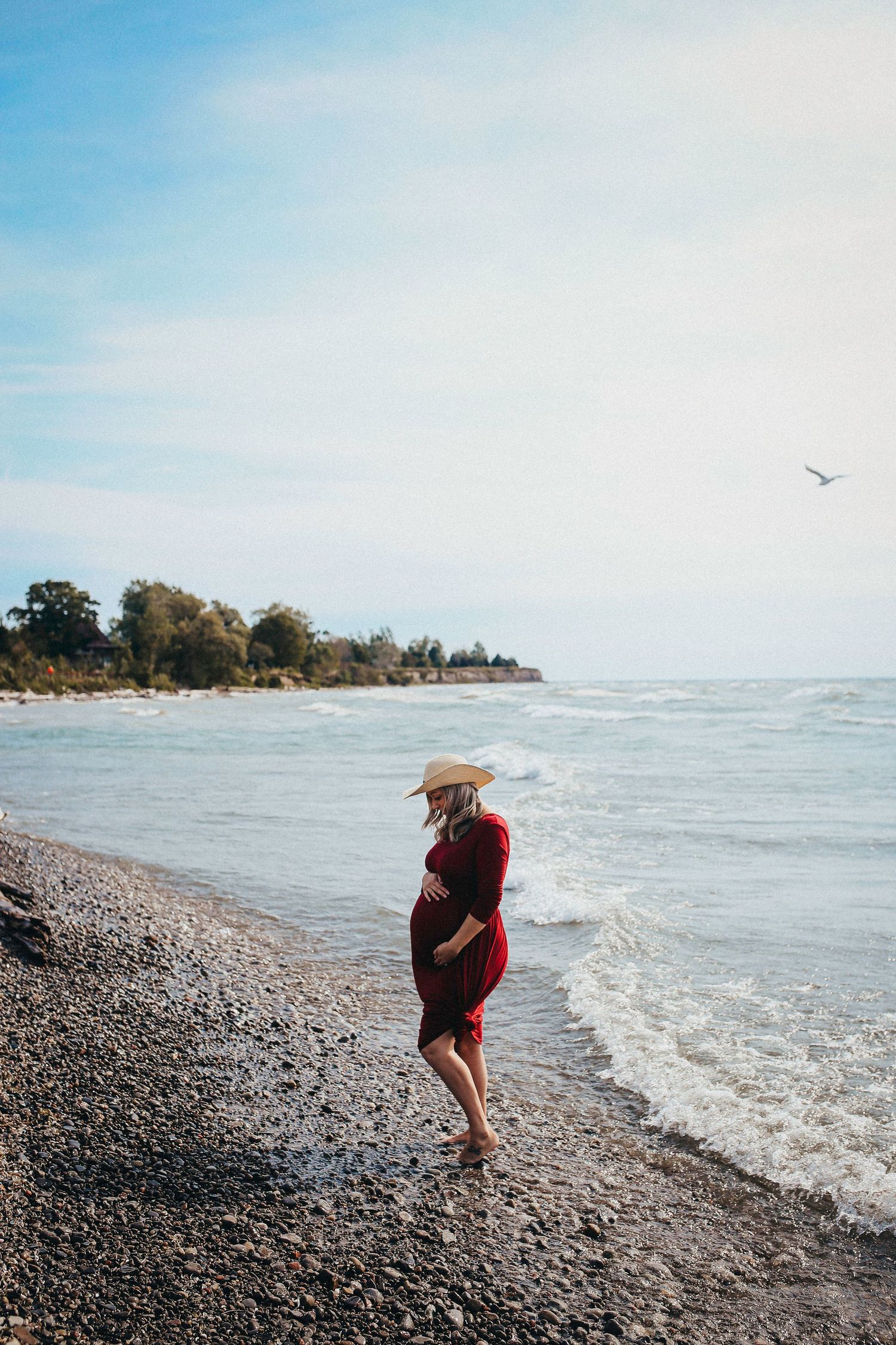 Woman in burgundy dress walking along a rocky beach shoreline on a bright day.