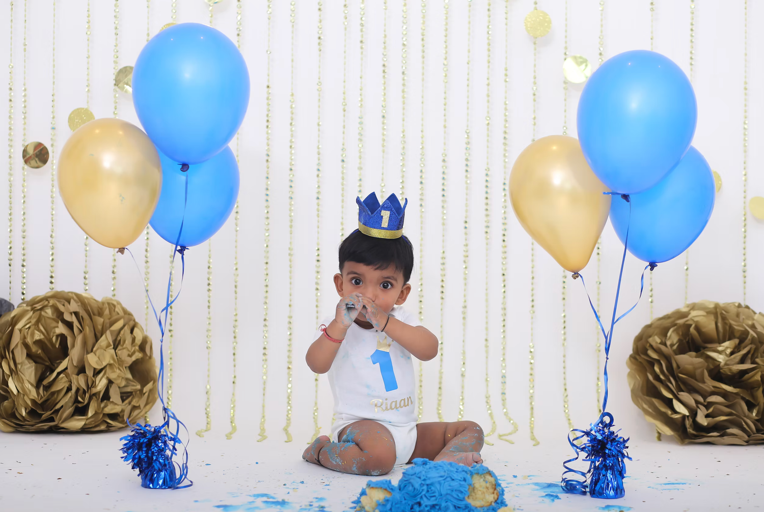 First birthday celebration setup with blue and gold balloons against white backdrop.