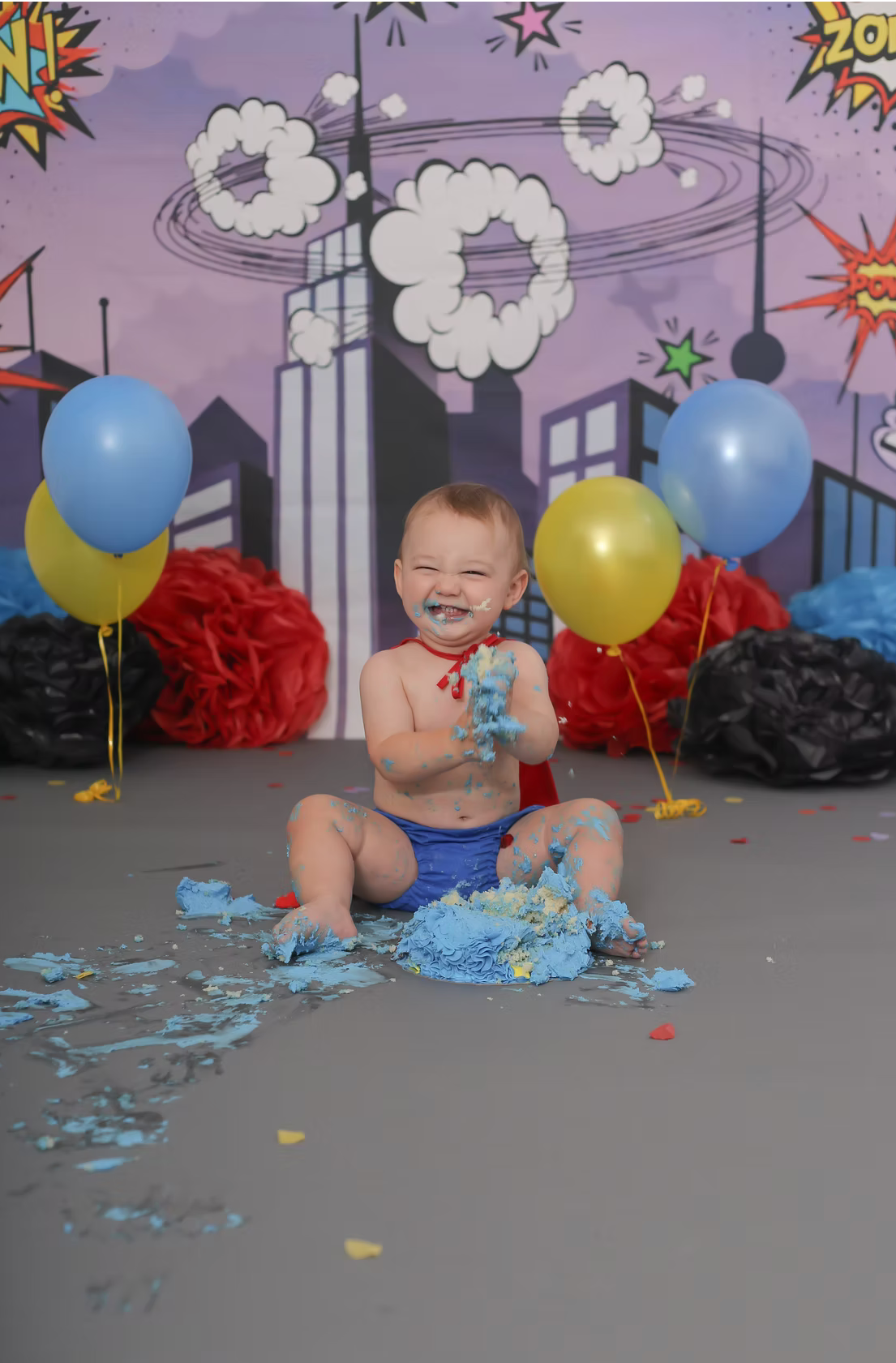 Superhero themed cake smash setup with blue frosting and colorful balloons against comic book backdrop.