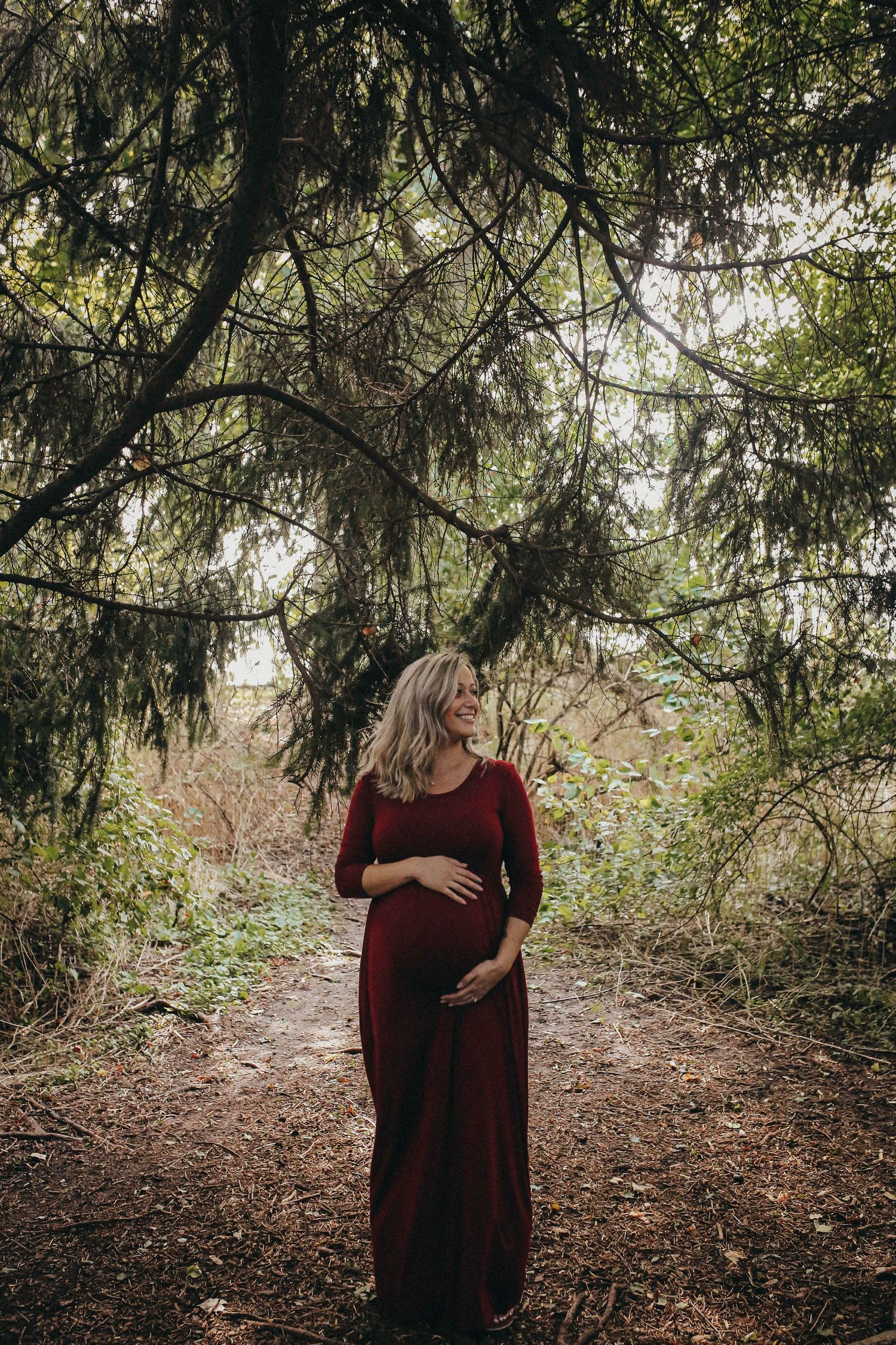 A person in a burgundy dress stands under evergreen trees in a forest setting.
