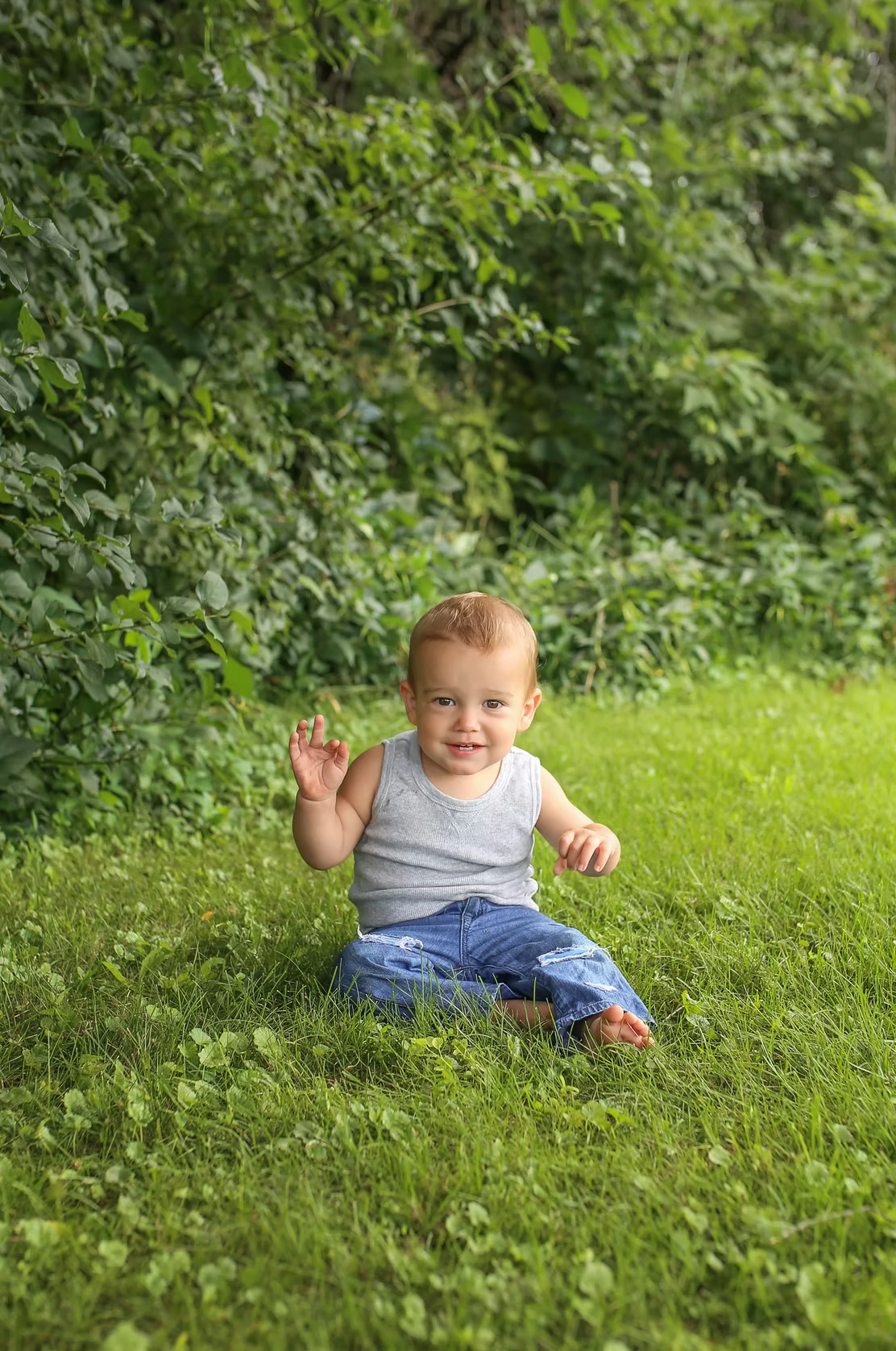 Small child sitting in grass waving with natural greenery in background.