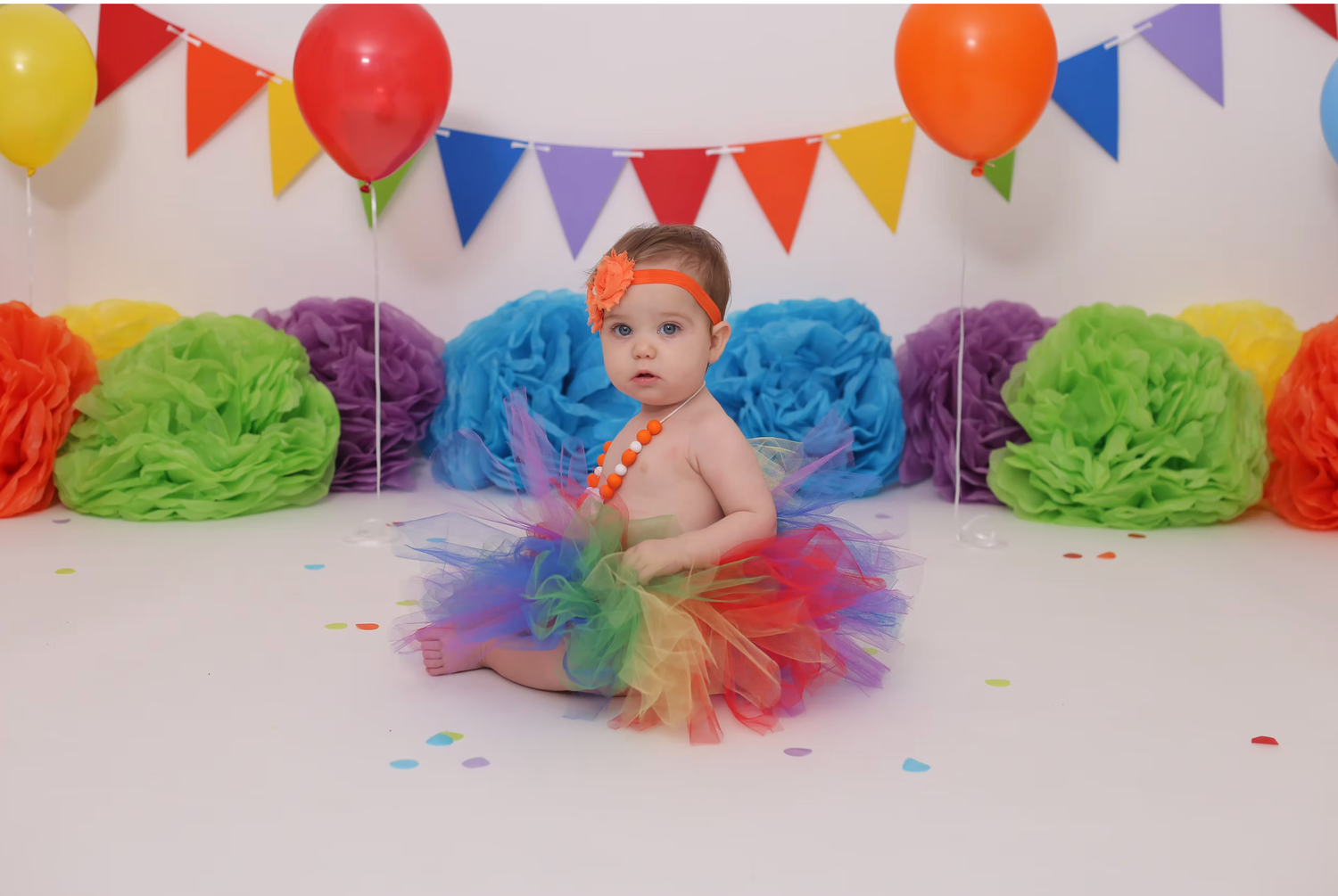 Colorful birthday party setup with rainbow tissue poms and festive bunting banners against white backdrop.