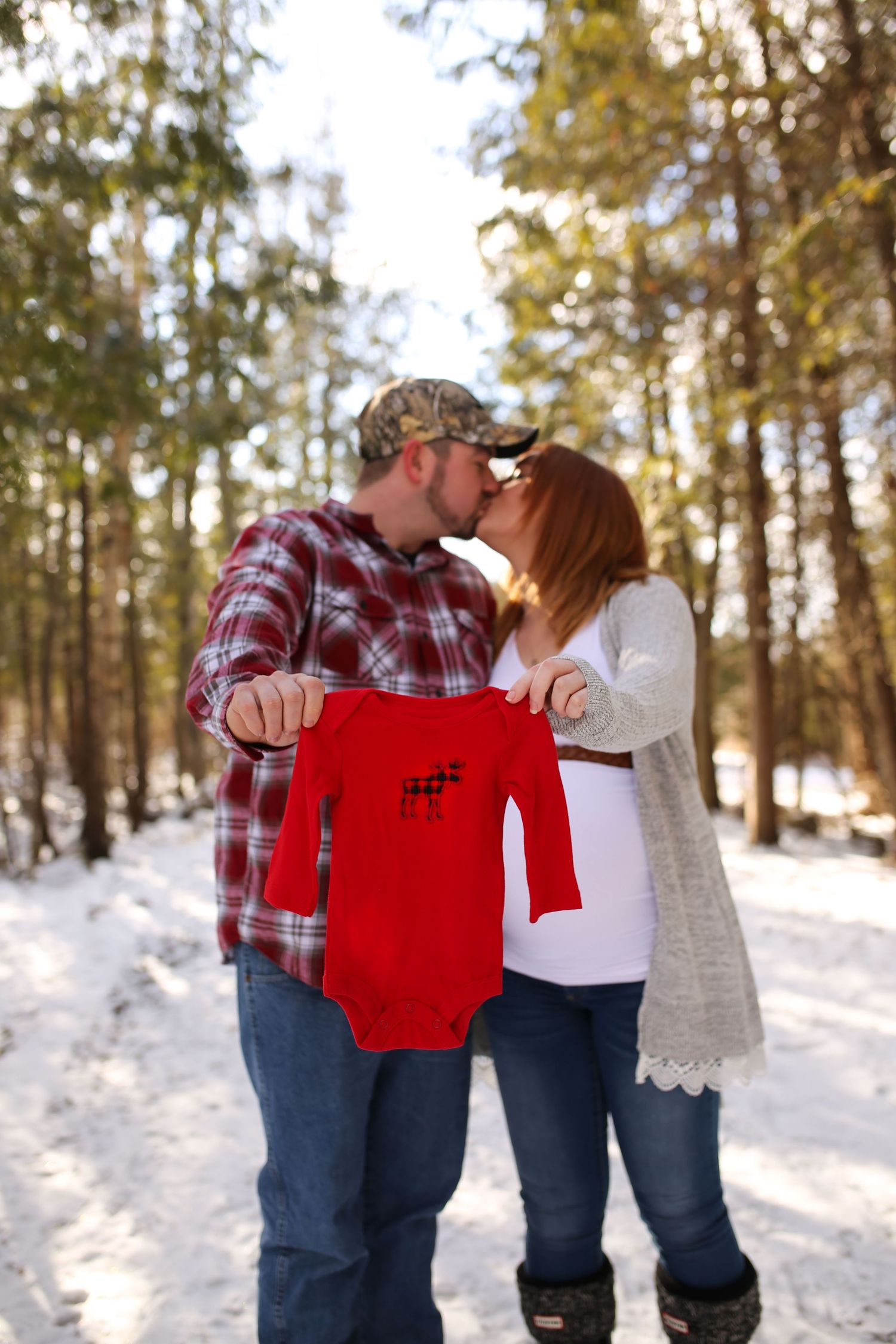 A couple holds up a tiny red baby outfit in a snowy winter forest scene.