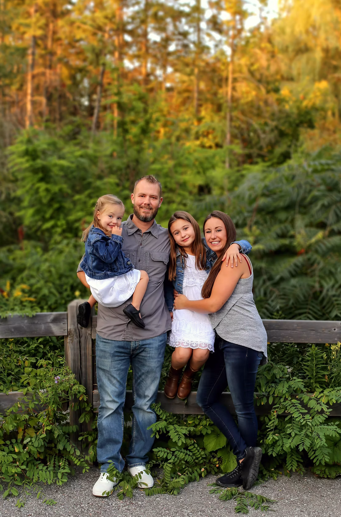 Family sitting together on a wooden bench surrounded by lush green ferns at sunset.