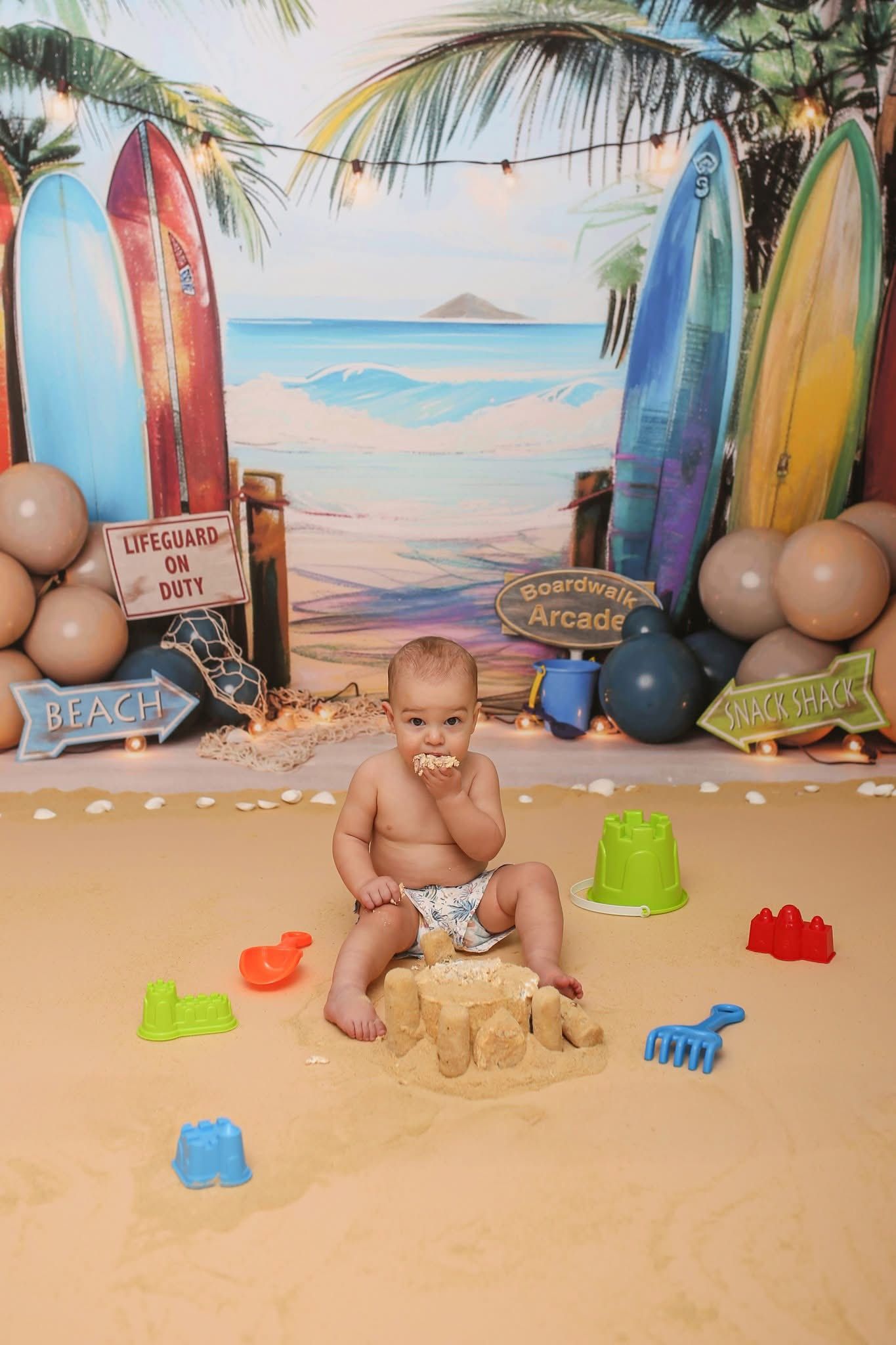 A beach-themed photo setup with surfboards and sand features a baby playing with colorful beach toys.