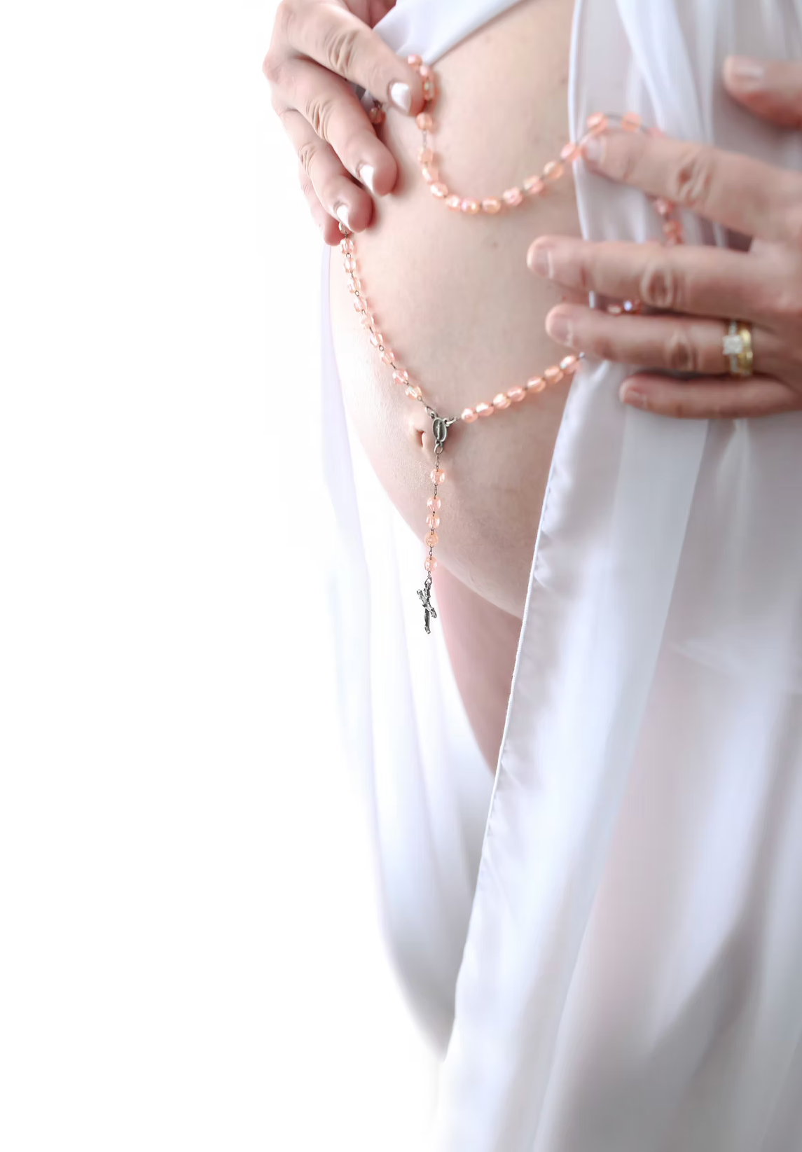 Hands hold a delicate pink rosary against a white fabric background in soft lighting.