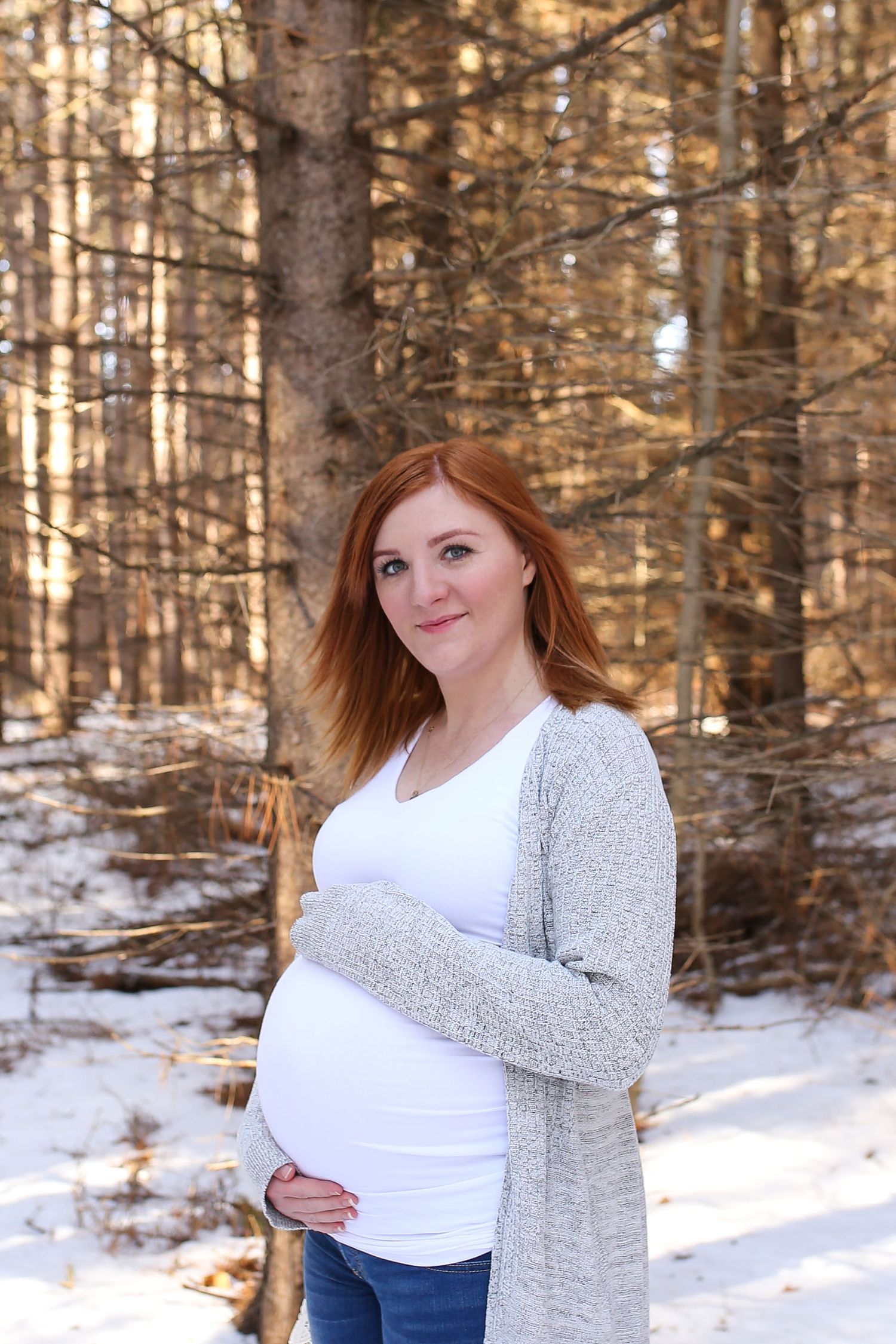 A winter maternity session in a snowy forest with sunlight streaming through the trees.