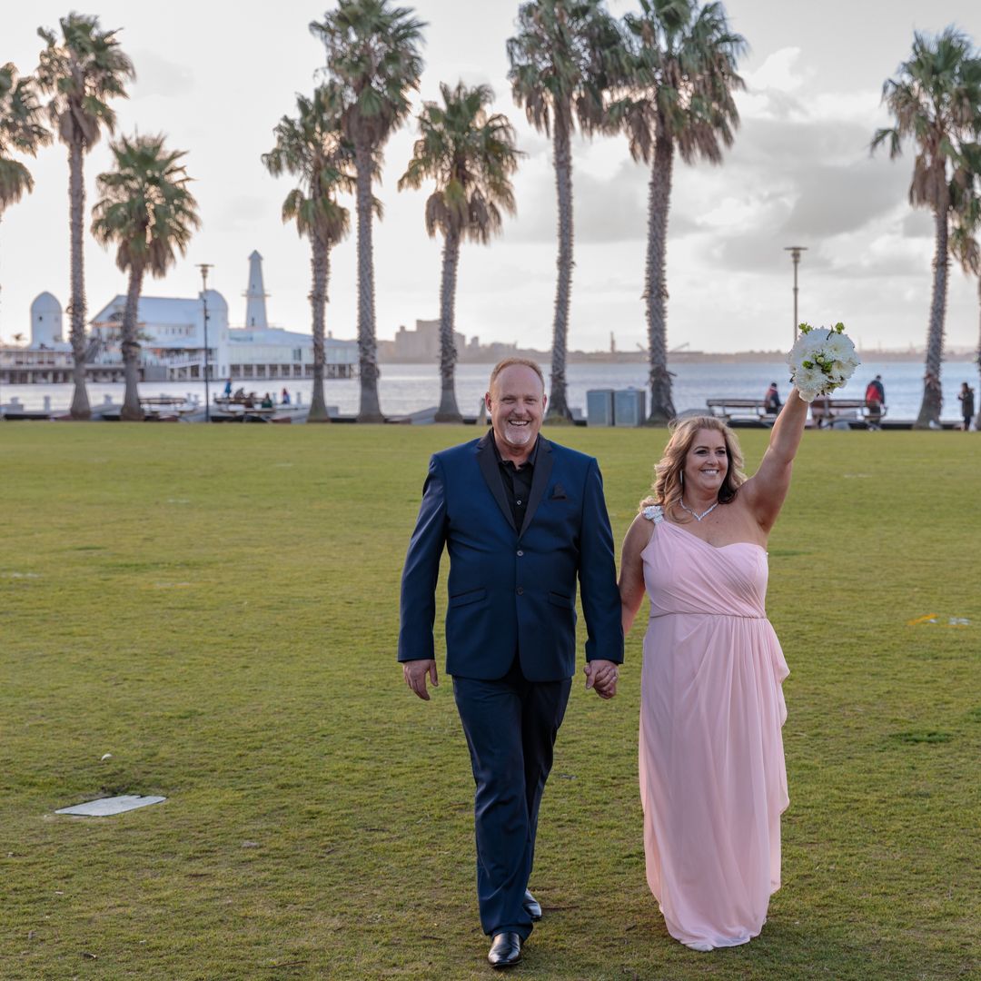 Bride and groom walking at Geelong Waterfront, Victoria. Bride has bouquet held in the air in celebration.