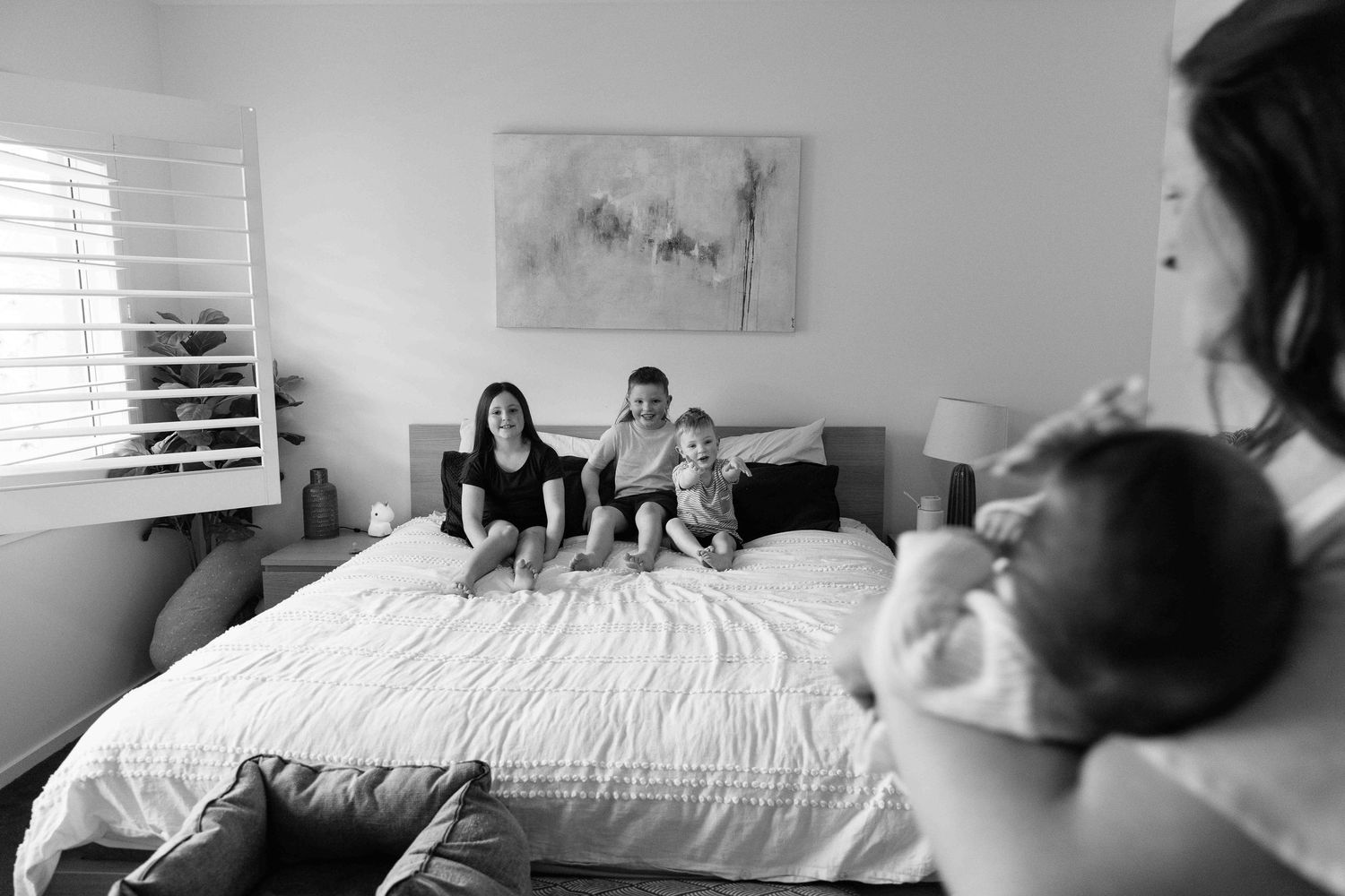 Mum walks past with baby while 3 siblings wait on the bed.  Leigh Ball Documentary Family Photographer