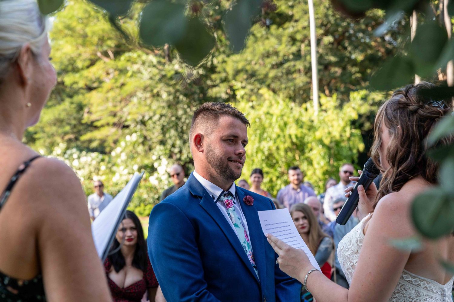 Bride reading wedding vows to groom at outdoor ceremony with friends and family looking on
