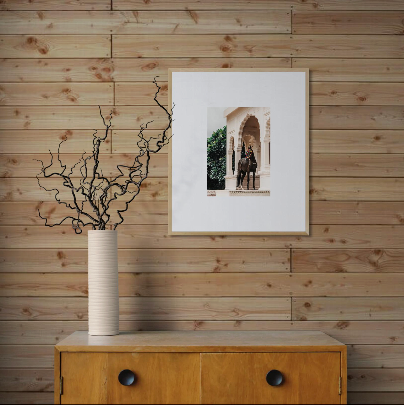 A framed architectural photo hangs on a wood-paneled wall above a vintage dresser with a white textured vase holding bare branches.